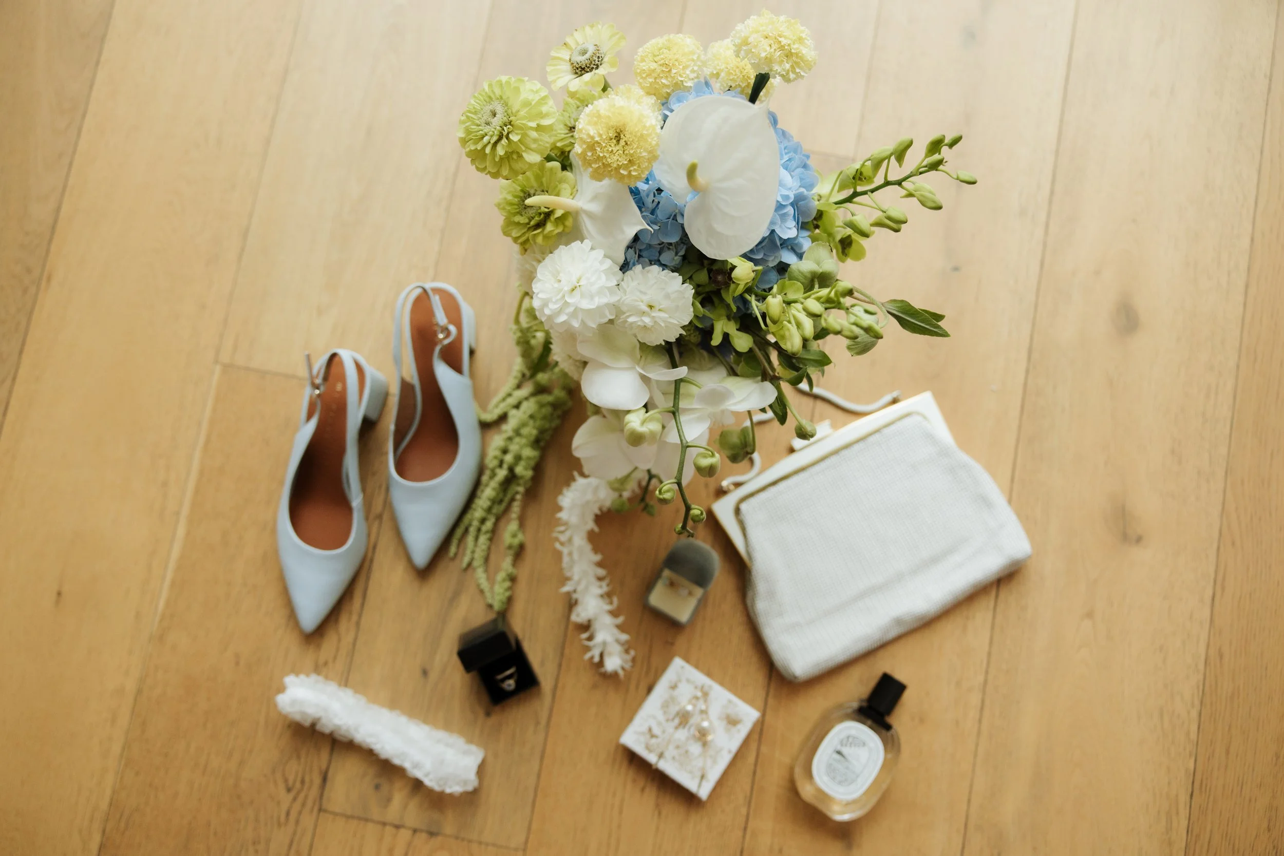 Arrangement of flowers, including white anthuriums, yellow and white hortensia, and other small flowers, placed on a wooden floor alongside a pair of light blue and brown high heels, a small white purse, and assorted jewelry and perfume bottles.