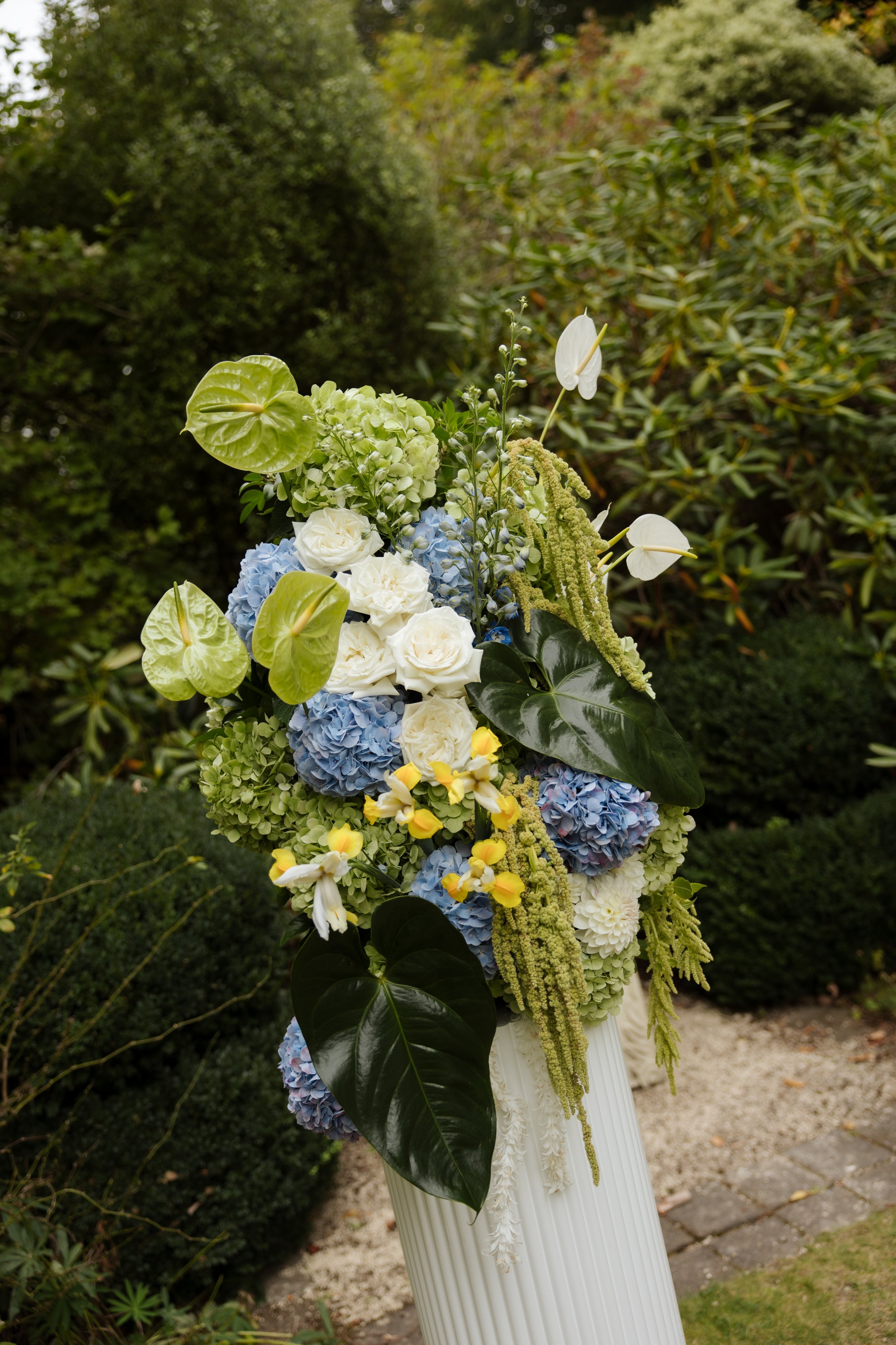 A floral arrangement with white roses, blue hydrangeas, white anthurium, and various green leaves on a white stand outdoors.