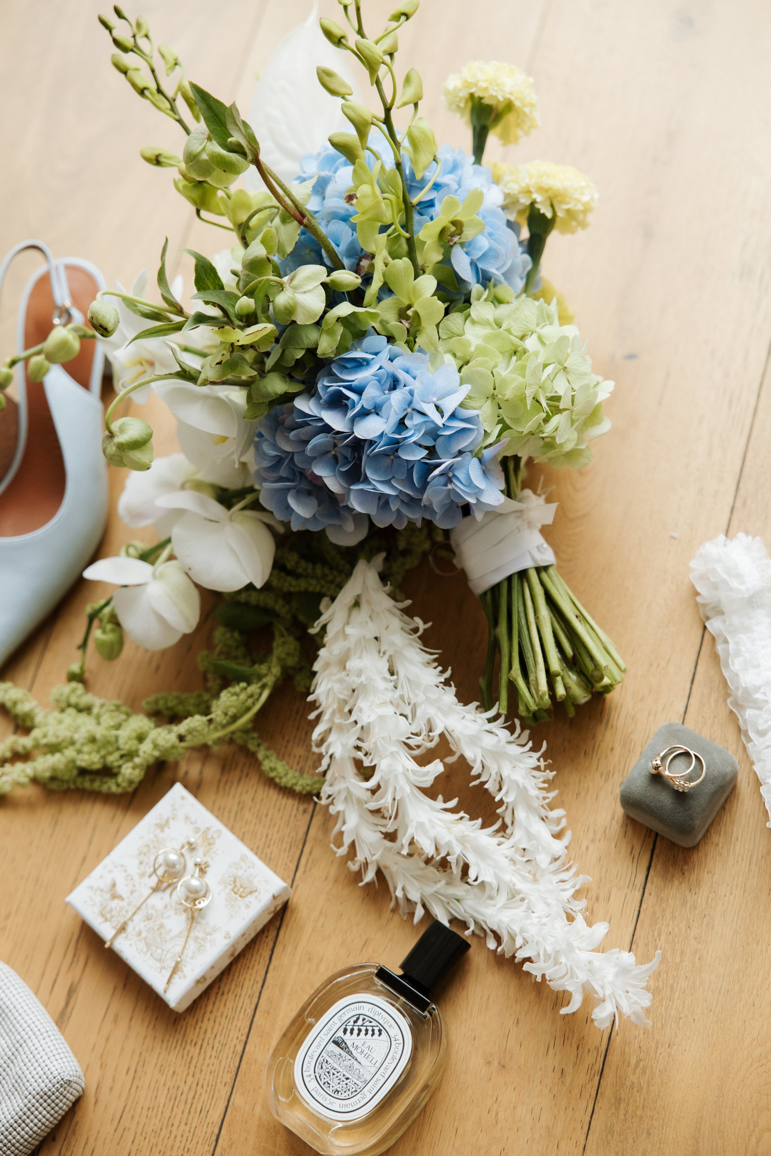 A bouquet of flowers including blue and yellow hydrangeas, white nerine, and greenery on a wooden surface. Nearby, there is a pair of earrings in a decorative box, a bottle of perfume, a gray ring box with a ring, a pair of white shoes, and jewelry.