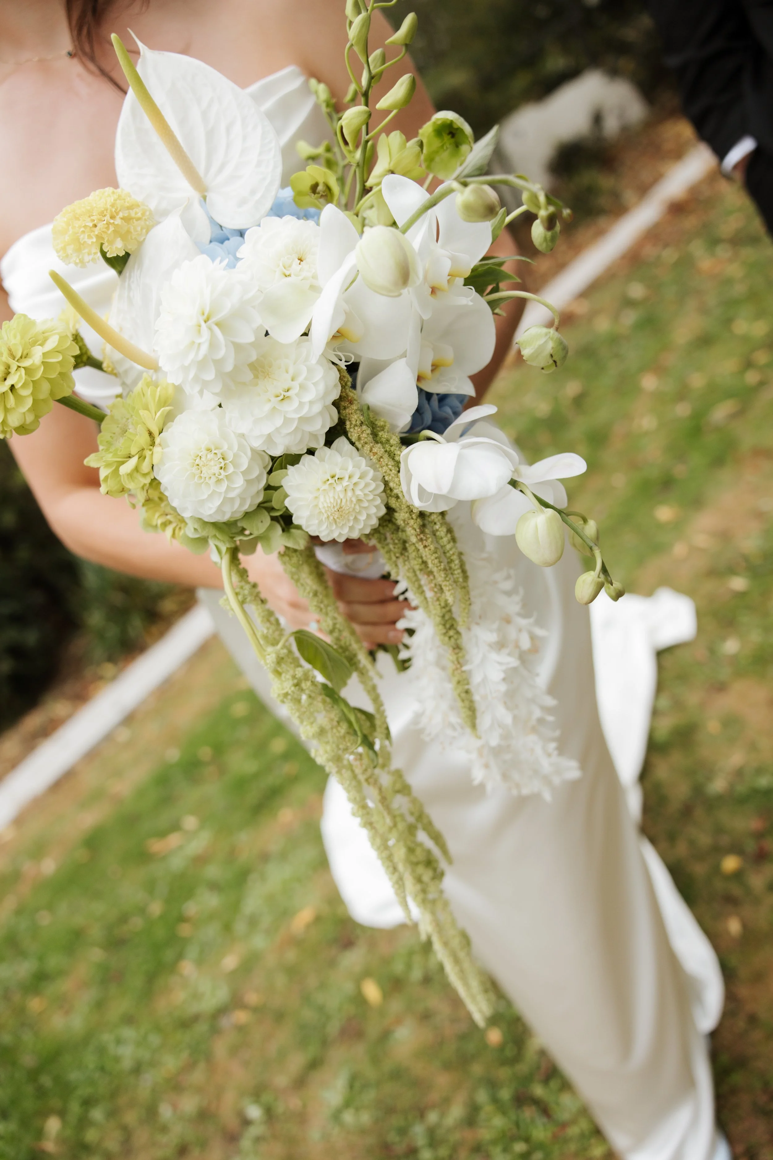 A bride holding a bouquet of white, cream, and green flowers outdoors with grass and fallen leaves in the background.