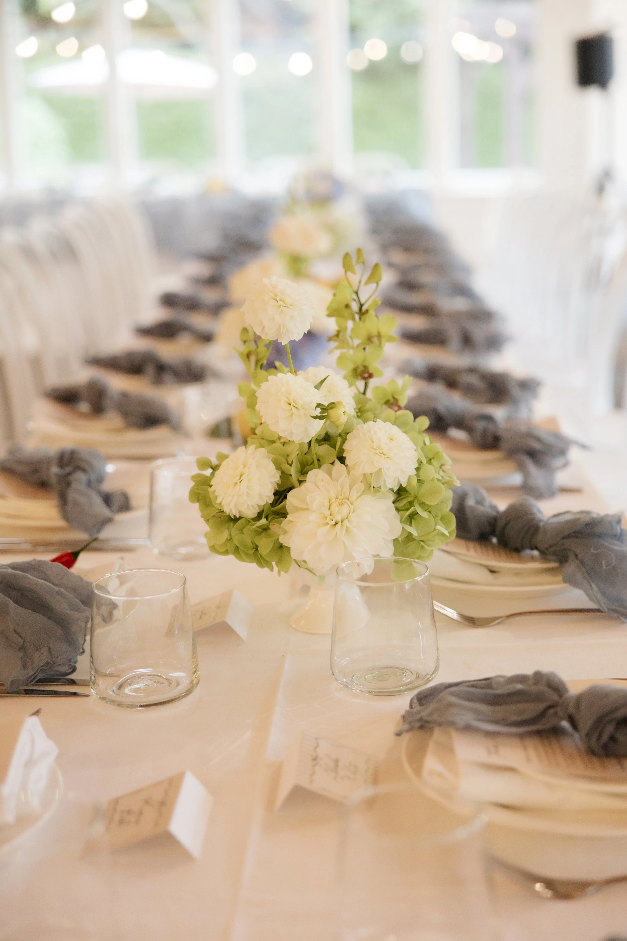 A long dining table set for a meal with a floral centerpiece of white and light green flowers. There are clear glass candle holders and place cards with printed text on the table. The table is decorated with gray napkins arranged around white plates.