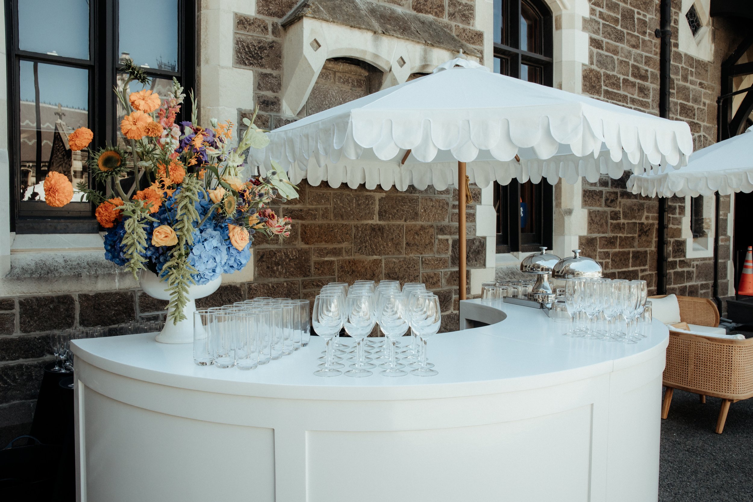 A round white table set up for a social event, featuring multiple empty wine glasses, a large floral arrangement with orange and purple flowers, and white umbrellas providing shade, all against a backdrop of a stone building with windows.