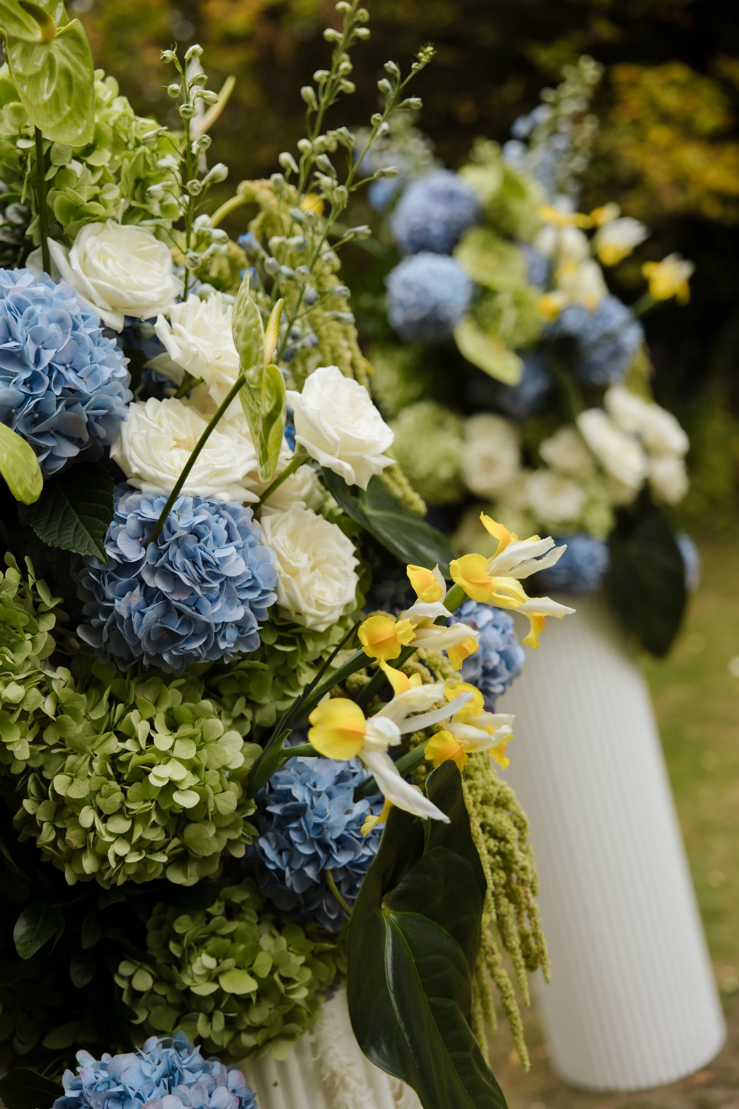 A floral arrangement with white roses, blue hydrangeas, yellow and white iris, green hydrangeas, and green foliage.