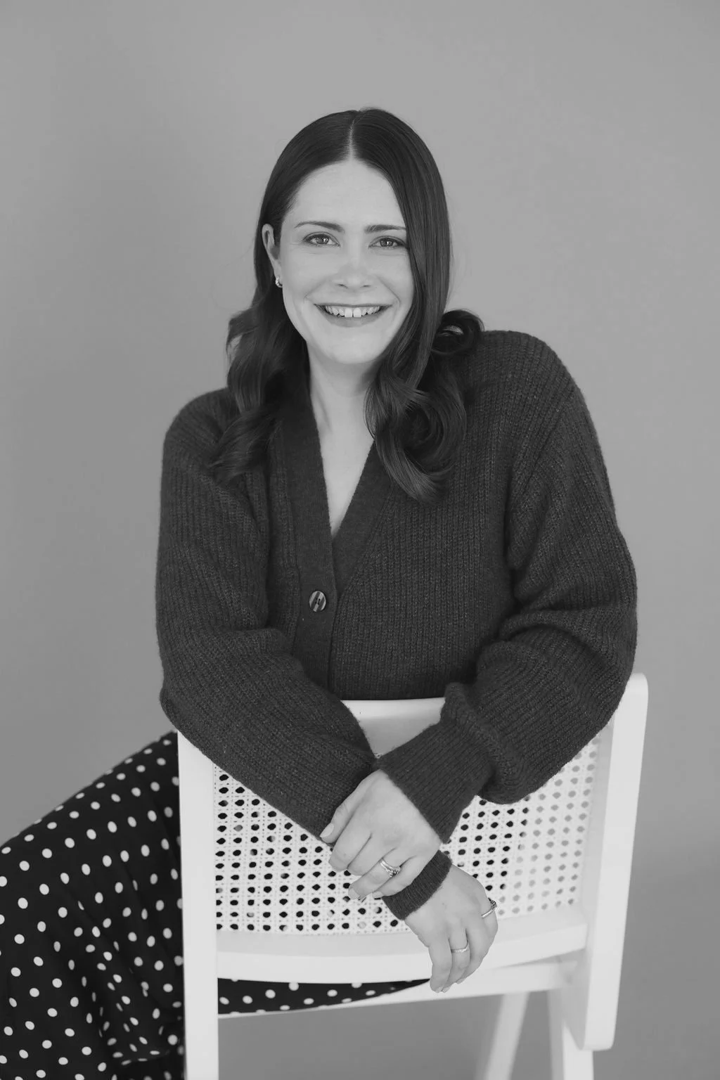 Black and white photo Emma from Linden Grove Floral with long, wavy hair sitting on a stool, smiling, wearing a dark sweater and polka dot pants, against a plain background.