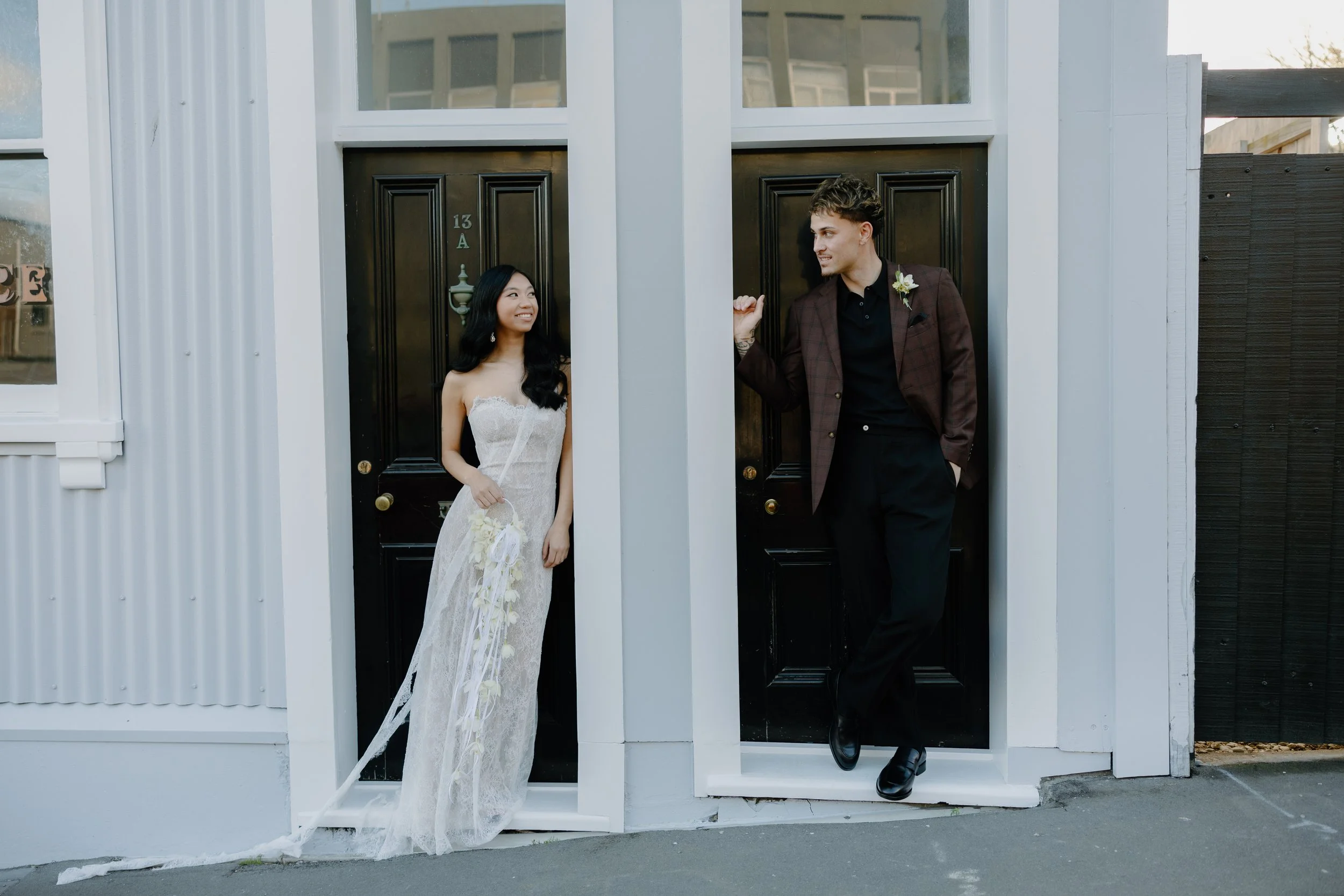 A young woman and a young man stand in front of a black door with the number 13A. The woman is in a wedding dress holding a bouquet, looking at the man, who is dressed in a suit with a boutonniere, leaning on the doorframe and looking at the woman.