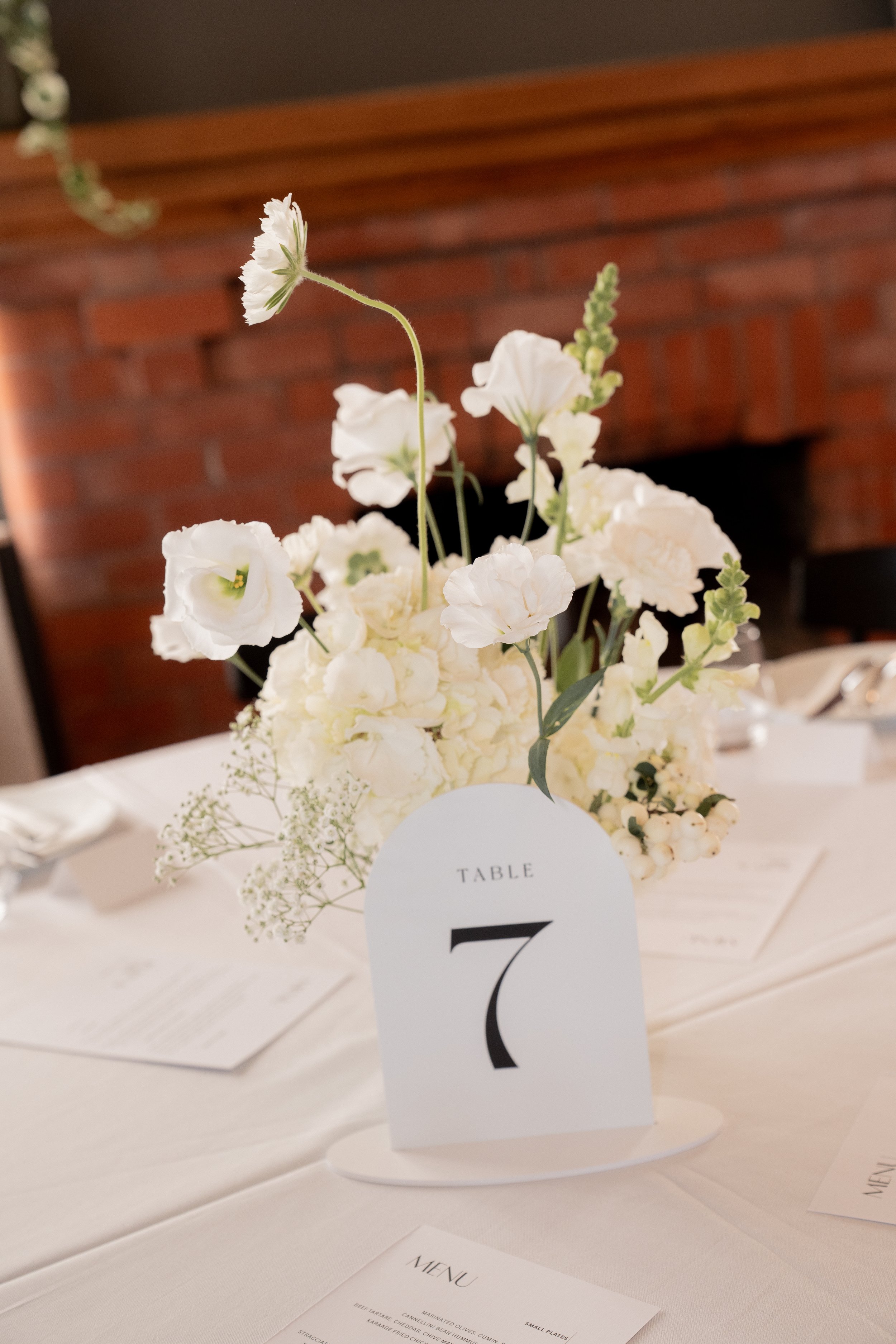 A flower arrangement with white and cream-colored flowers on a table, with a table number card showing the number 7, in a formal setting with menus and a white tablecloth.