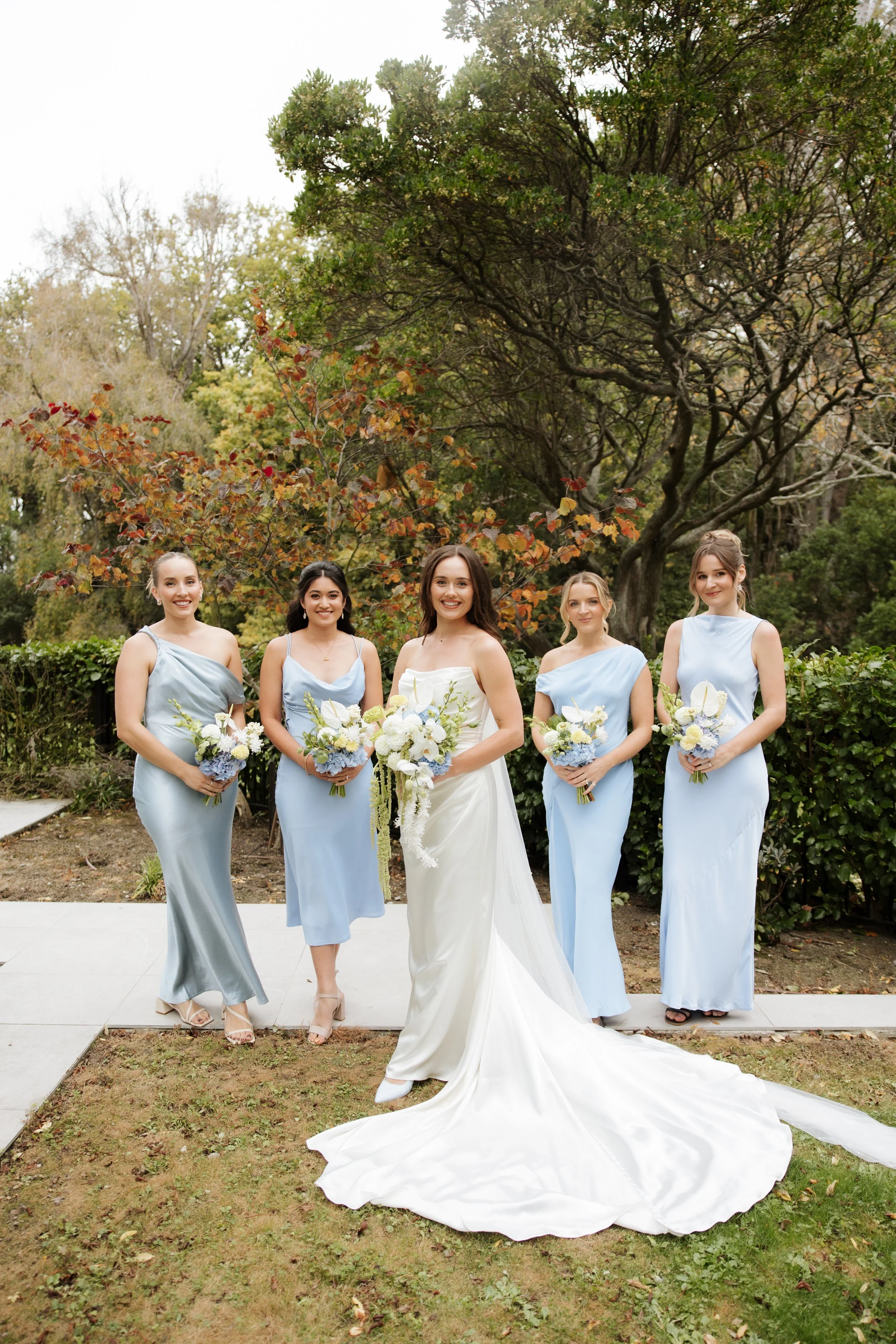 Five women standing outdoors in formal attire, with four in light blue dresses and one in a white wedding gown, holding bouquets of flowers, in front of trees with autumn leaves.