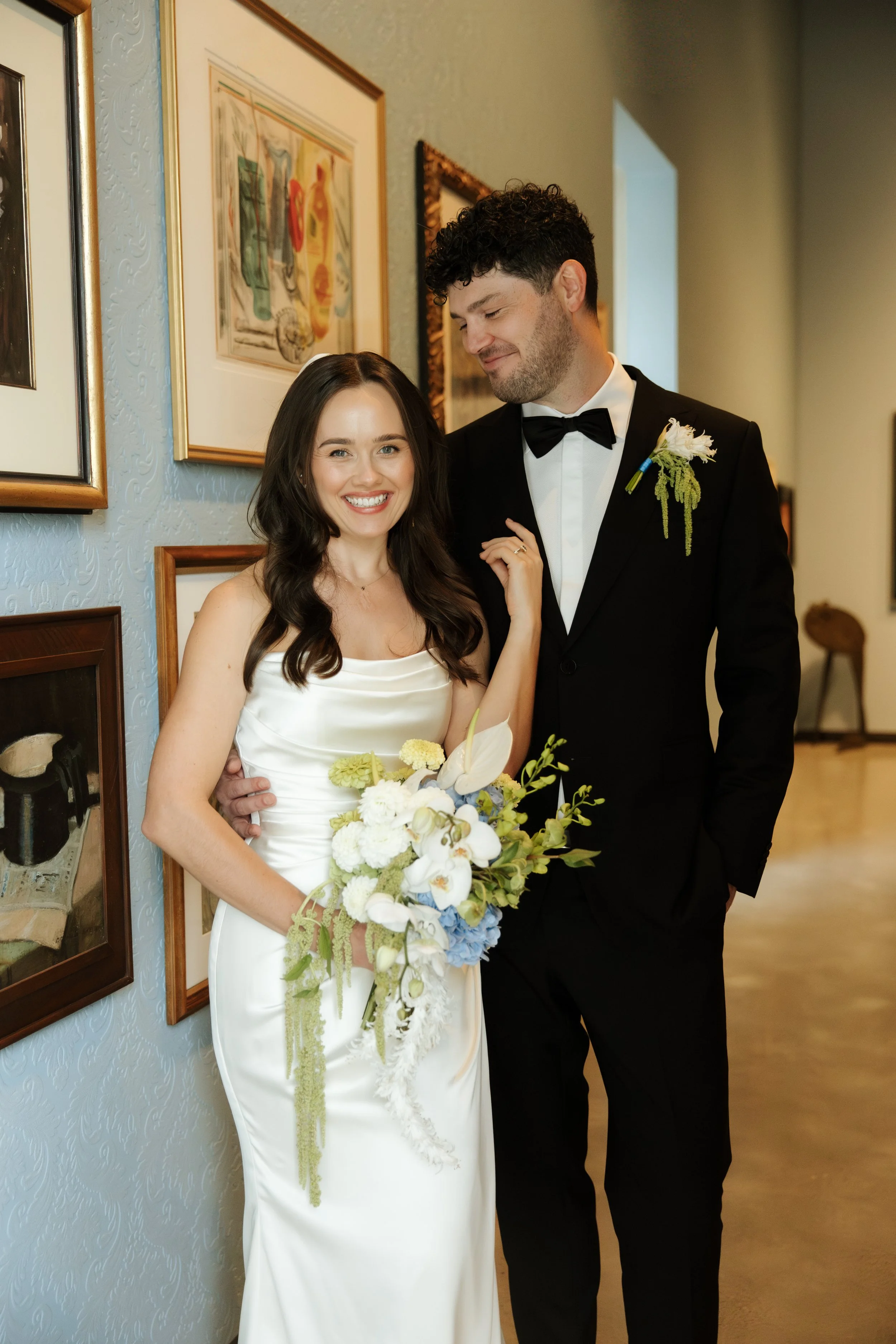 A smiling woman in a white wedding dress holding a large bouquet of white and blue flowers, standing next to a man in a black tuxedo with a boutonniere, both indoors near a wall with framed artwork.