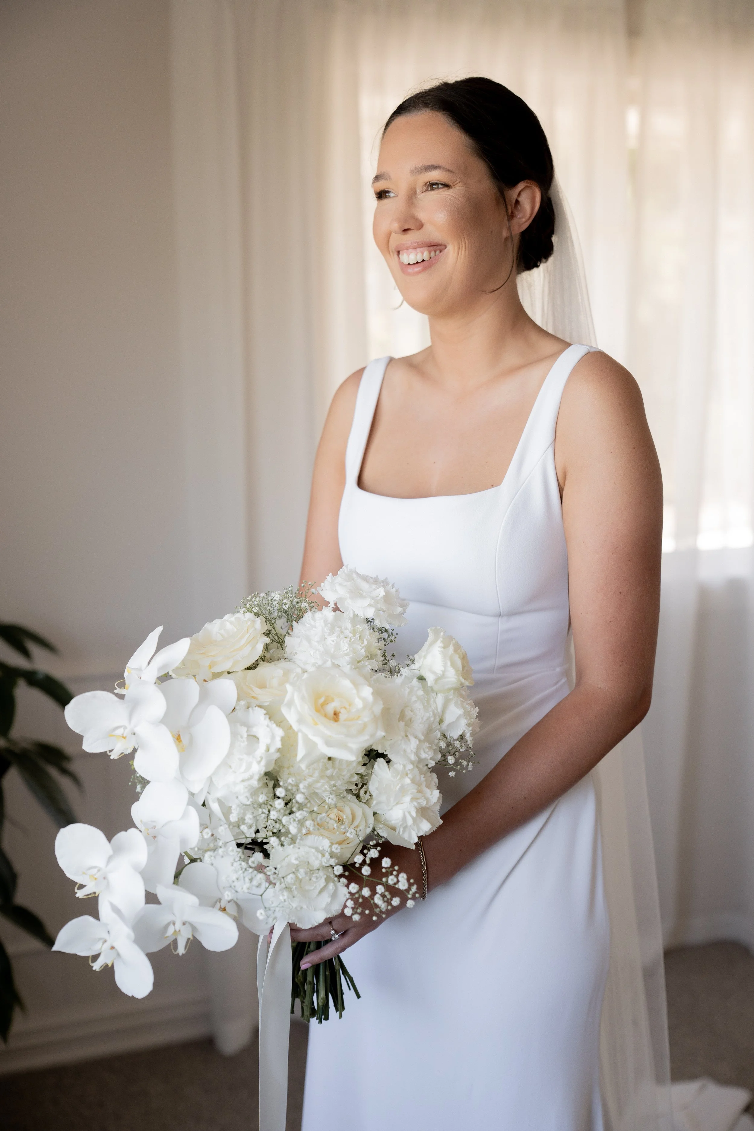 A smiling bride in a white wedding dress holding a bouquet of white flowers, including orchids and roses, standing in front of a light-colored curtain.