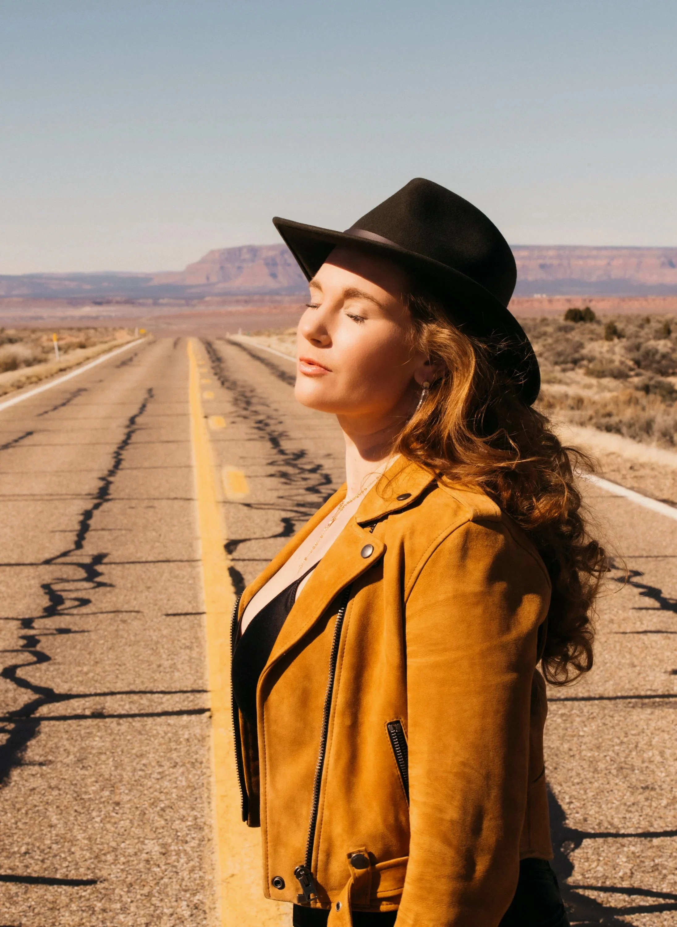 A woman with curly hair wearing a brown jacket and a black hat, standing on an empty desert road with mesas in the background, rocks, and sparse vegetation.
