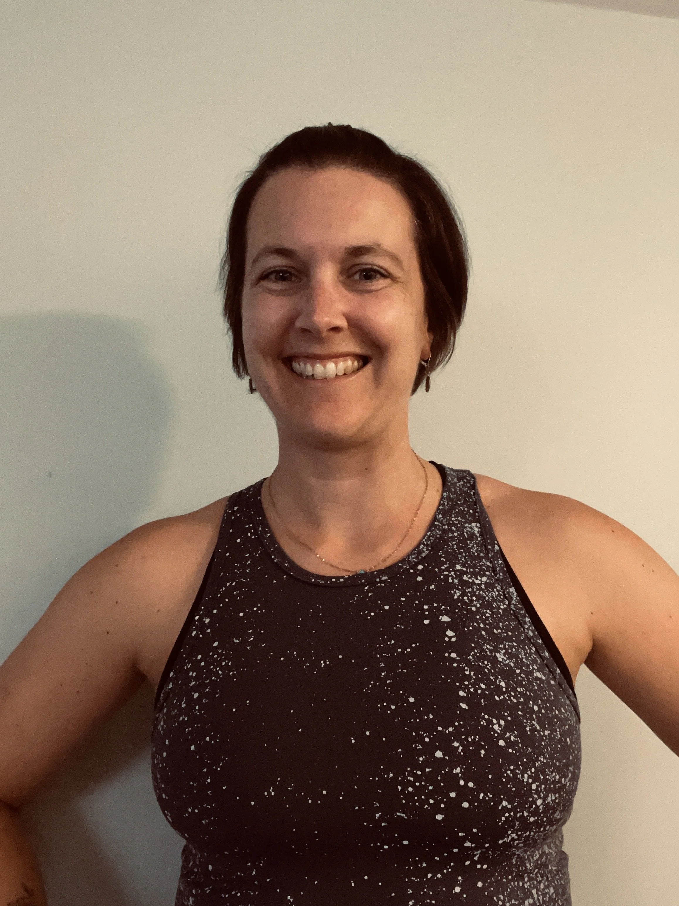A woman with short brown hair smiling, wearing a black sleeveless sports top with white speckled pattern, standing against a plain light-colored wall.