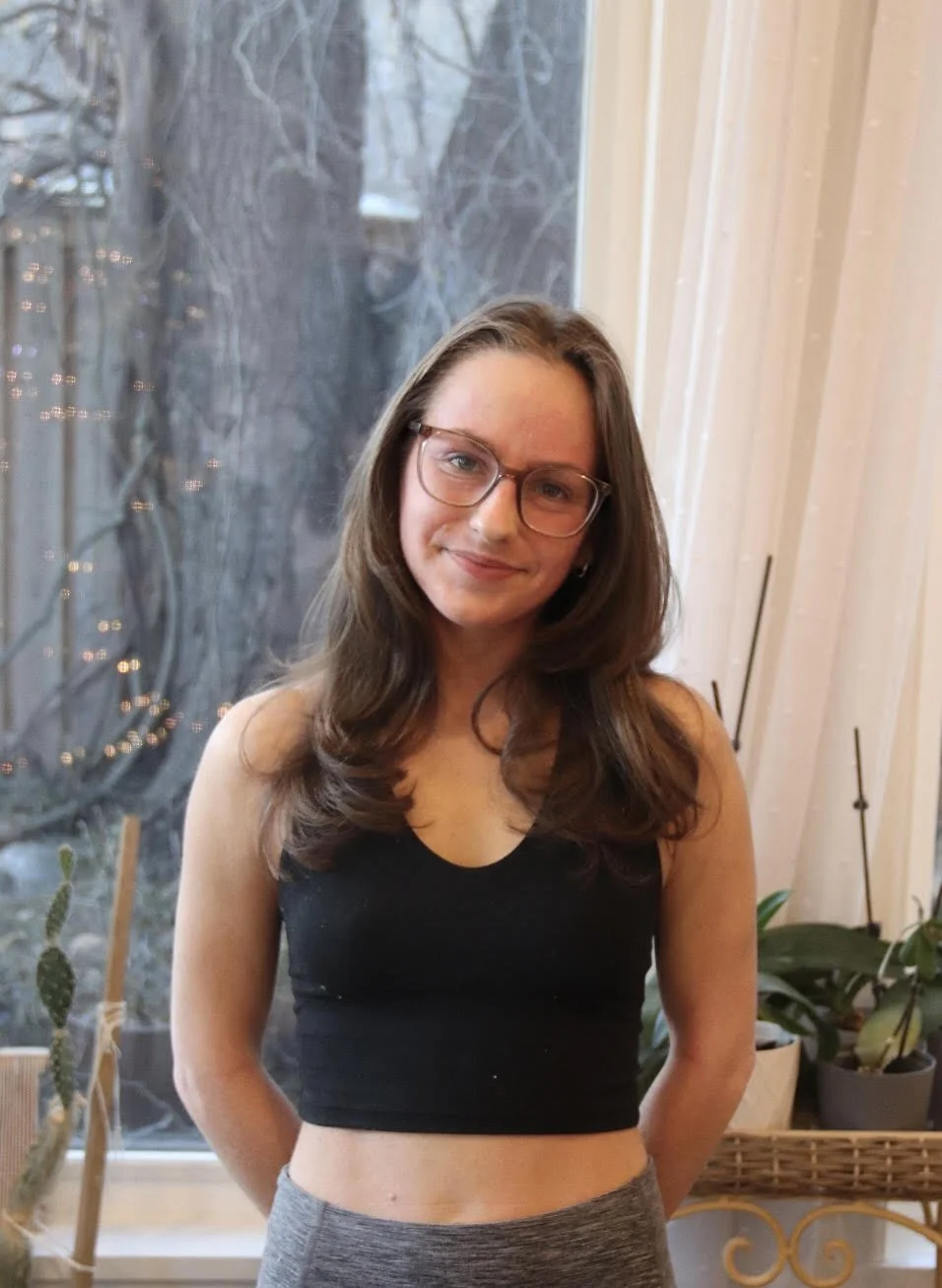 A woman with long brown hair, glasses, and a black crop top standing indoors near a window with plants and fairy lights in the background.