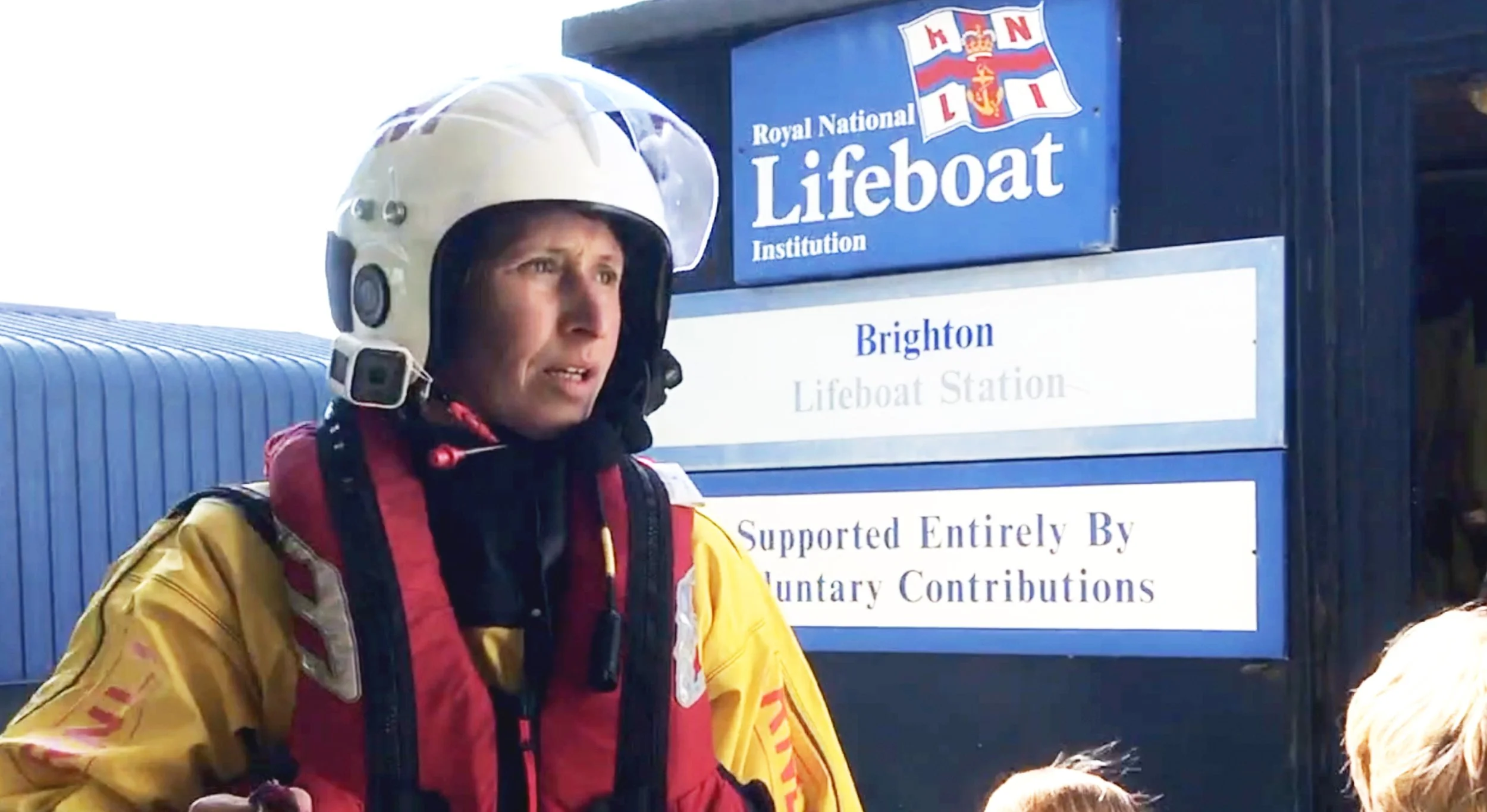 RNLI volunteer Irenka Wood stands in protective gear outside Brighton Lifeboat Station, a contemporary photograph highlighting volunteer rescue work, maritime humanitarianism and quiet British heroism. Mothership Adrift circumnavigators