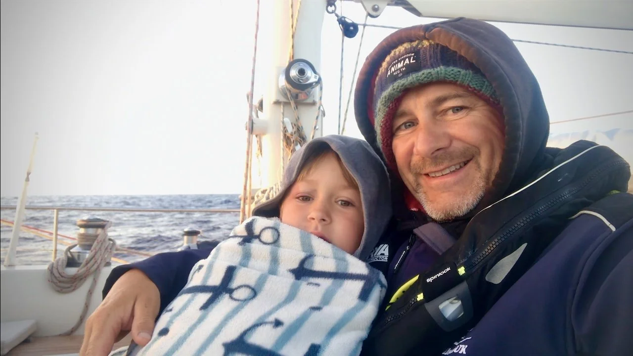 Father and son - Woody and Yewan on S/V Mothership cuddling up to keep warm in a sailboat cockpit
