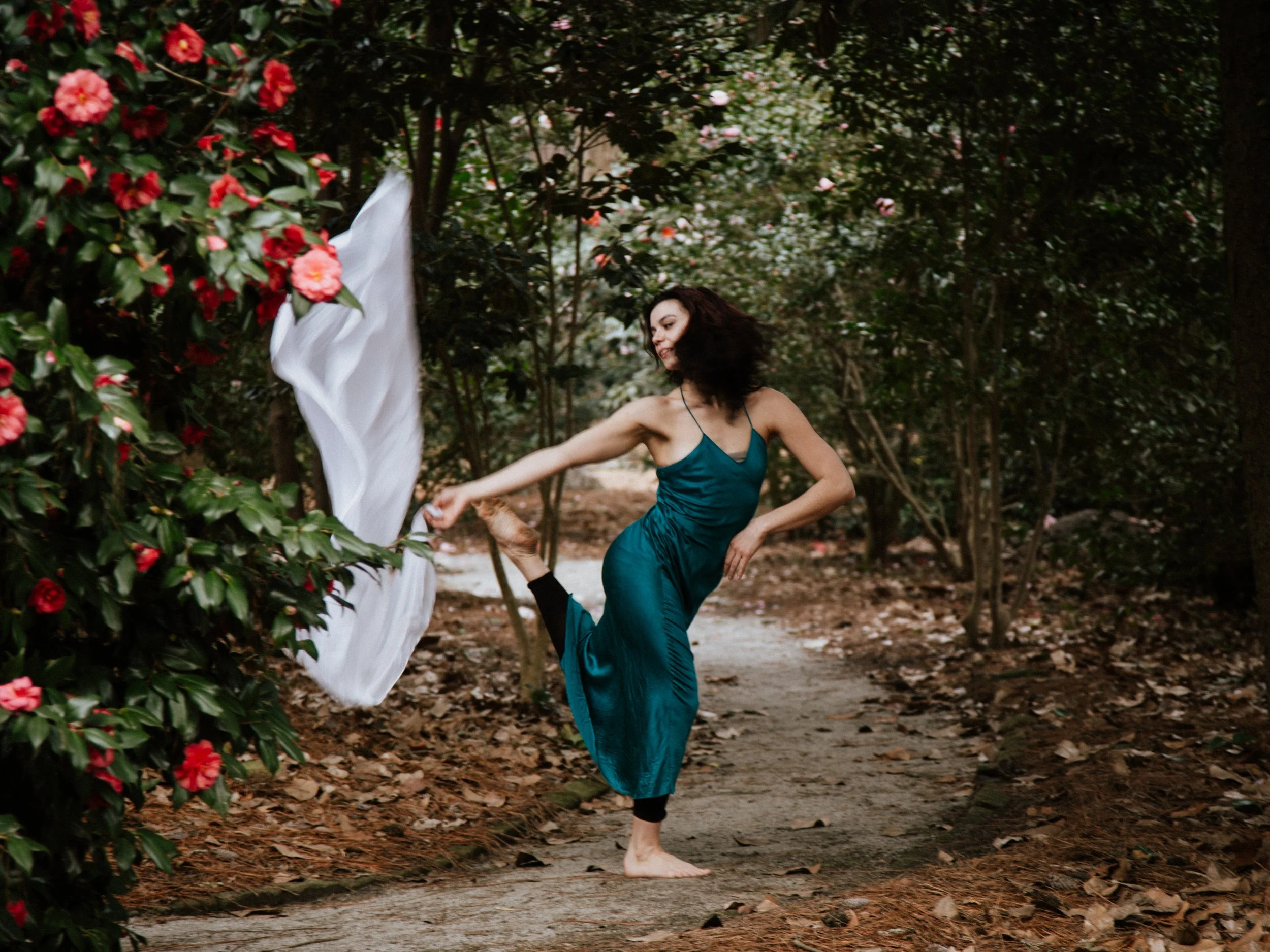 A woman in a teal satin dress stretches her leg behind her while walking barefoot on a wooded path lined with red and pink flowers.