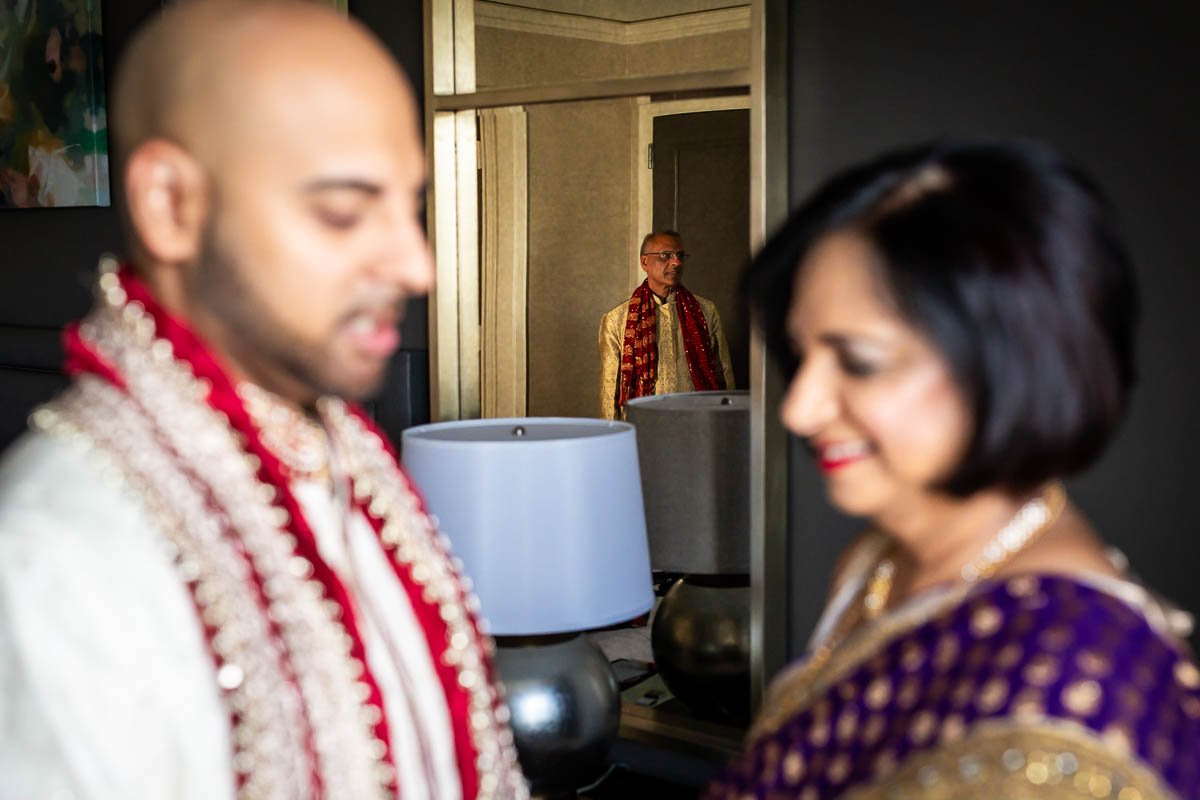  Dad looks on in mirror reflection as mom helps groom get ready before Indian wedding at Ritz-Carlton Tysons Corner 
