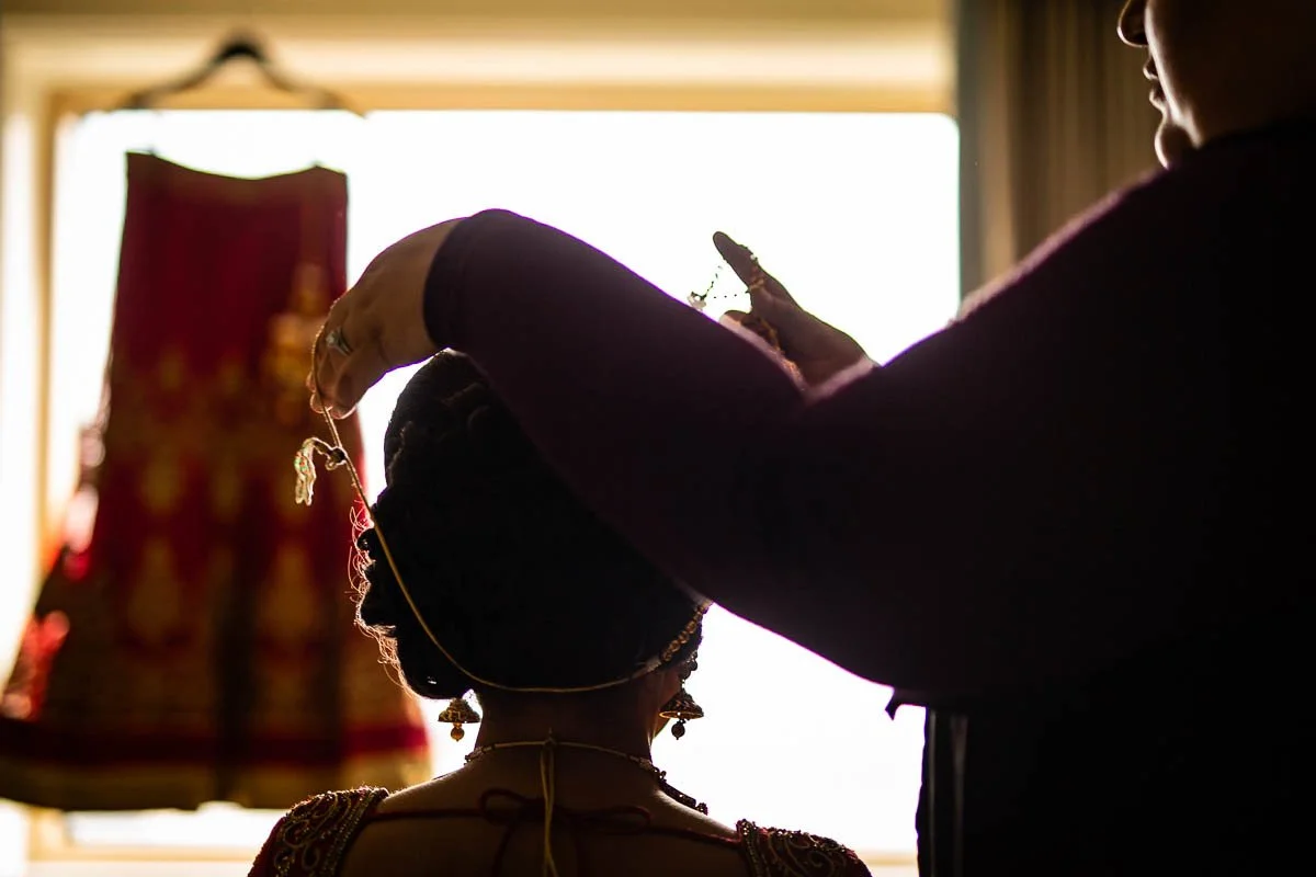 Mahwish Ali helping put on necklace on bride in front of bridal lehenga on hanger before Virginia Indian wedding