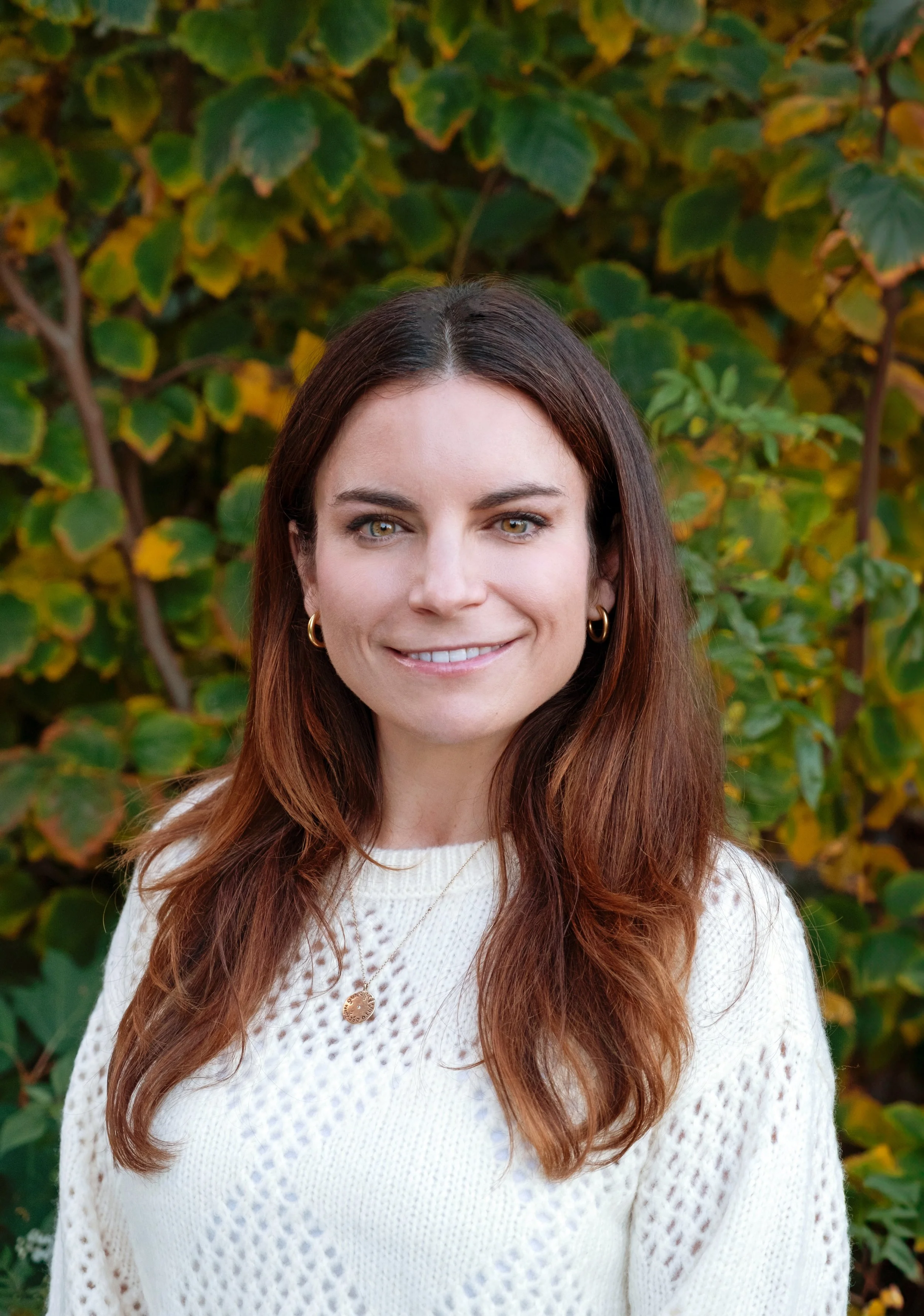 Portrait of a young woman with long brown hair, wearing a cream-colored knitted sweater, and gold hoop earrings, standing outdoors in front of green and yellow foliage.