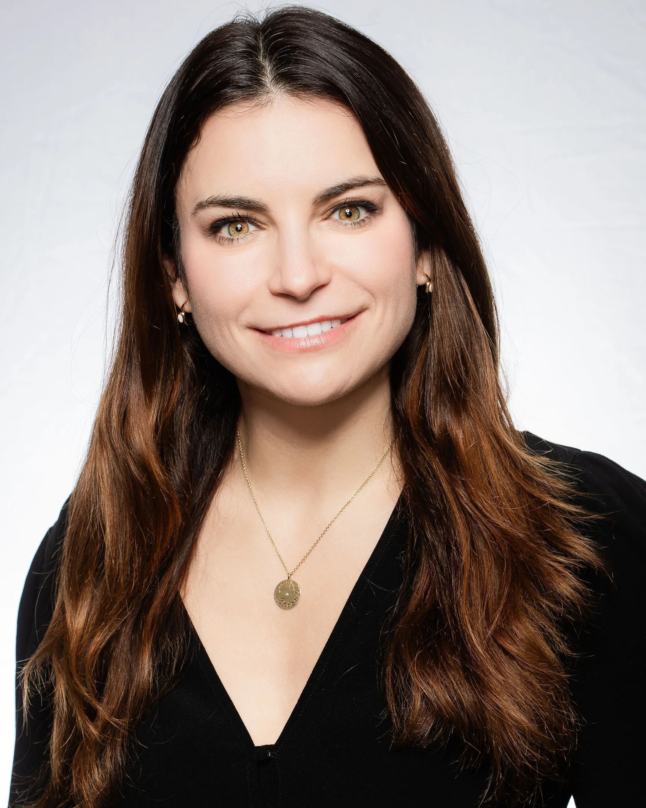 Close-up of a woman with long brown hair and gold eyes, smiling, wearing a black top and gold necklace.