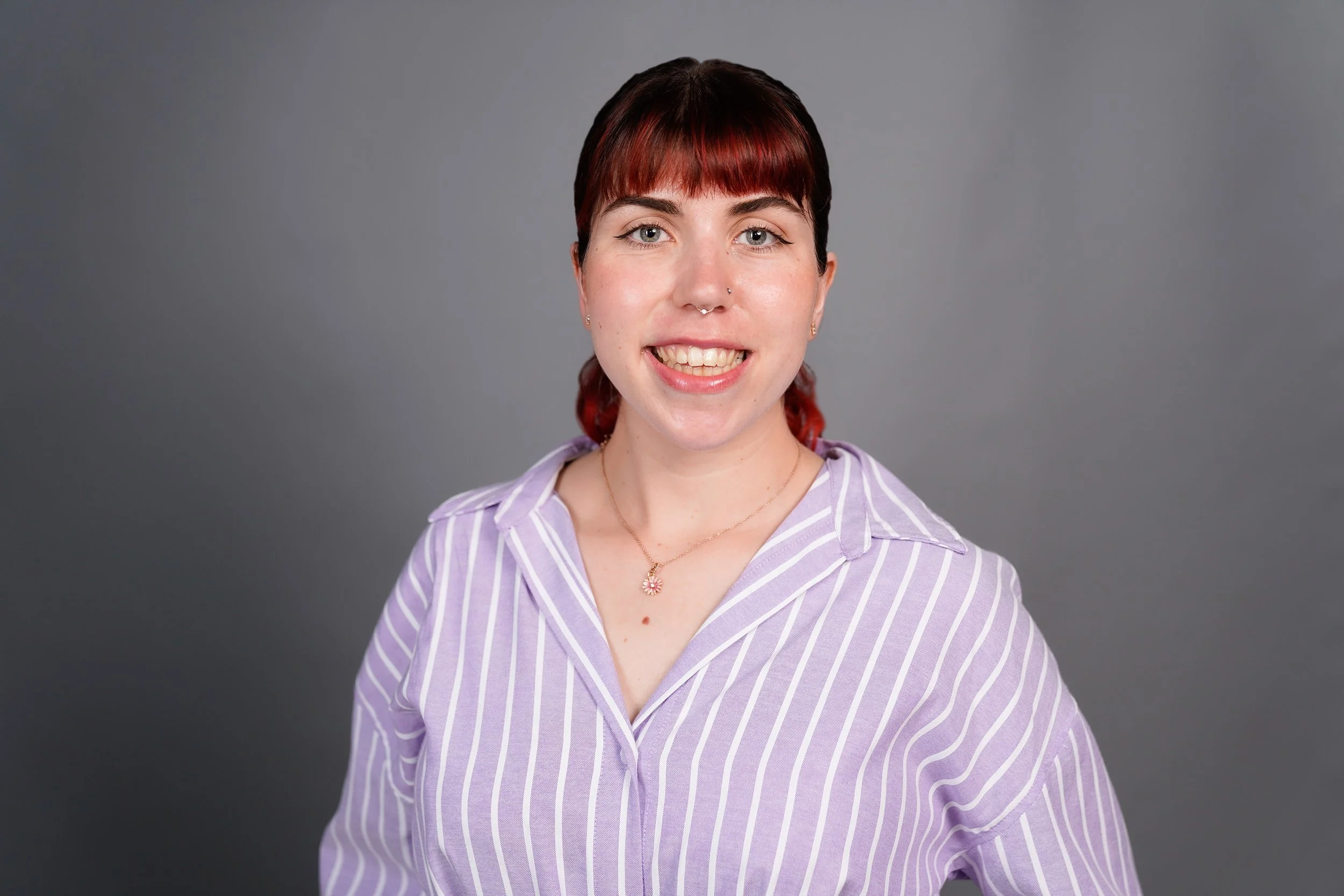 A woman with short dark red hair, light blue eyes, and a septum piercing smiling at the camera, wearing a purple and white striped shirt and a gold necklace with a pink flower pendant, standing against a plain gray background.