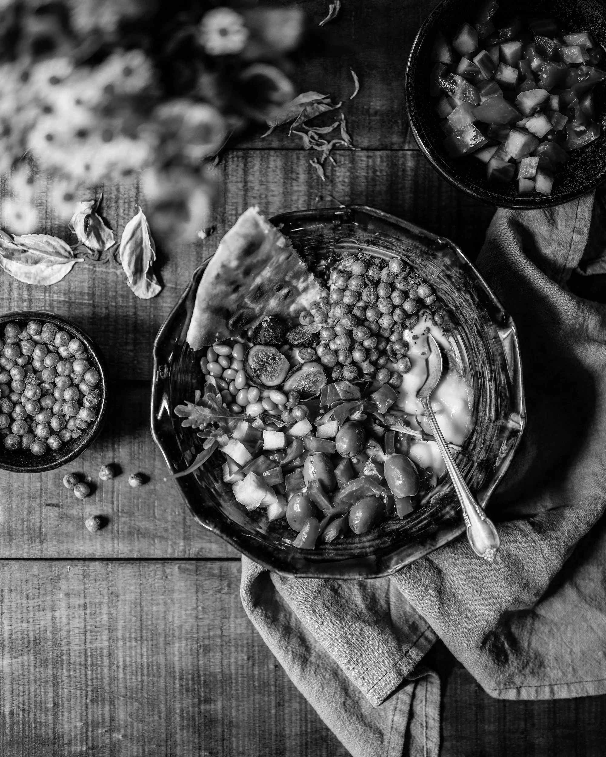 Black and white image of a salad in a bowl with a spoon, containing vegetables, grains, and a slice of watermelon, on a wooden table.