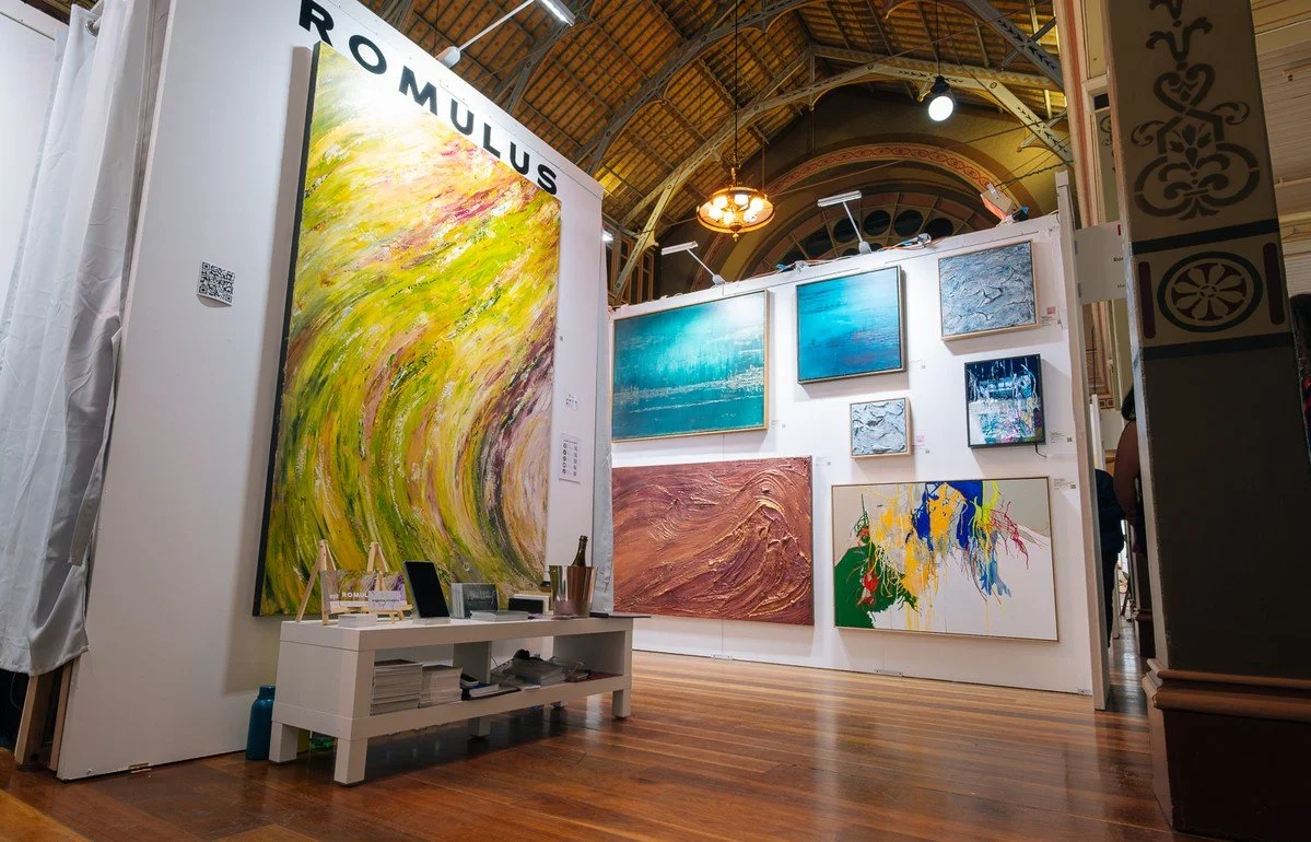 An art gallery showcasing multiple abstract paintings on white walls, including blue, green, and pink tones, with wooden flooring and a decorative arched ceiling.