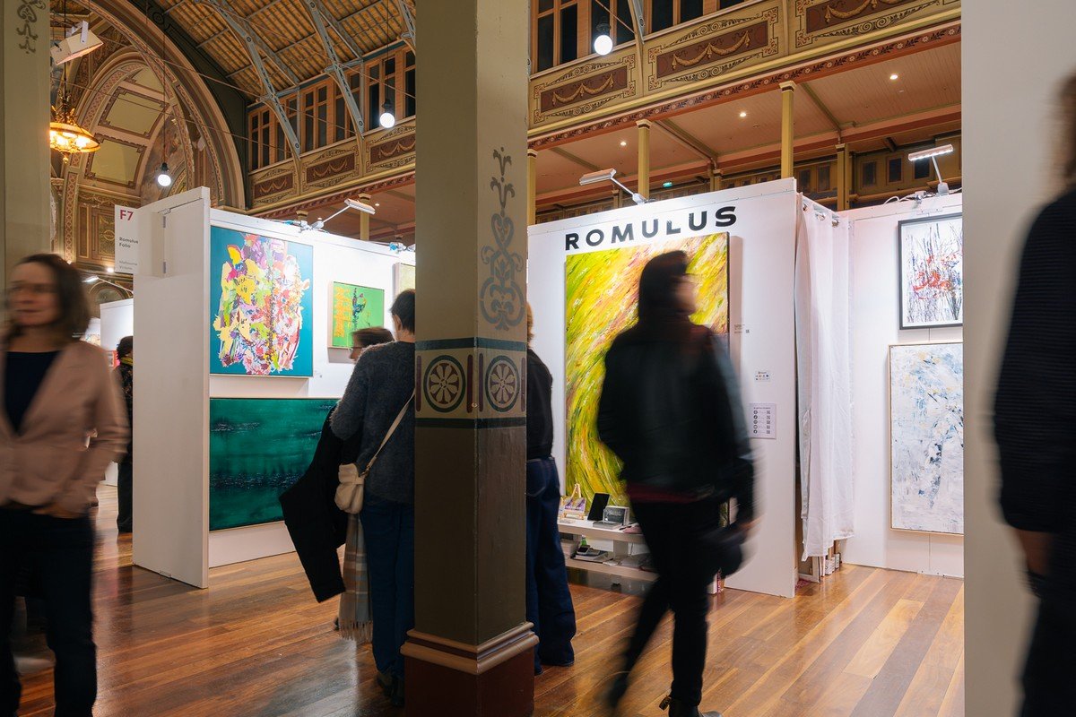 People walking through an art exhibition with various colorful paintings on display, in a grand hall with ornate architecture and high ceilings.