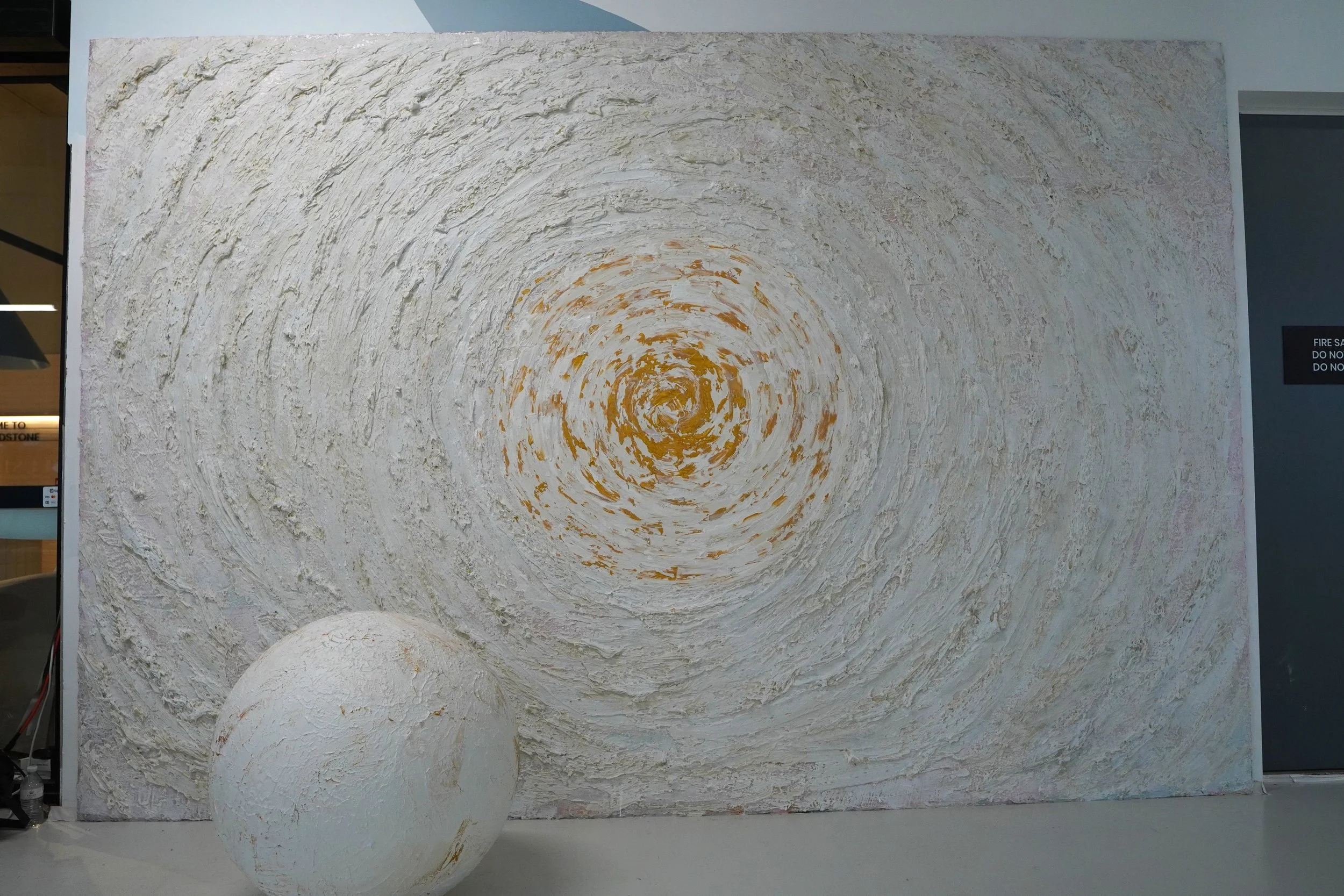 Large textured abstract painting with circular, swirl patterns in shades of white, cream, and hints of yellow, with a 3D spherical sculpture in front of it, also textured and painted in similar colors.