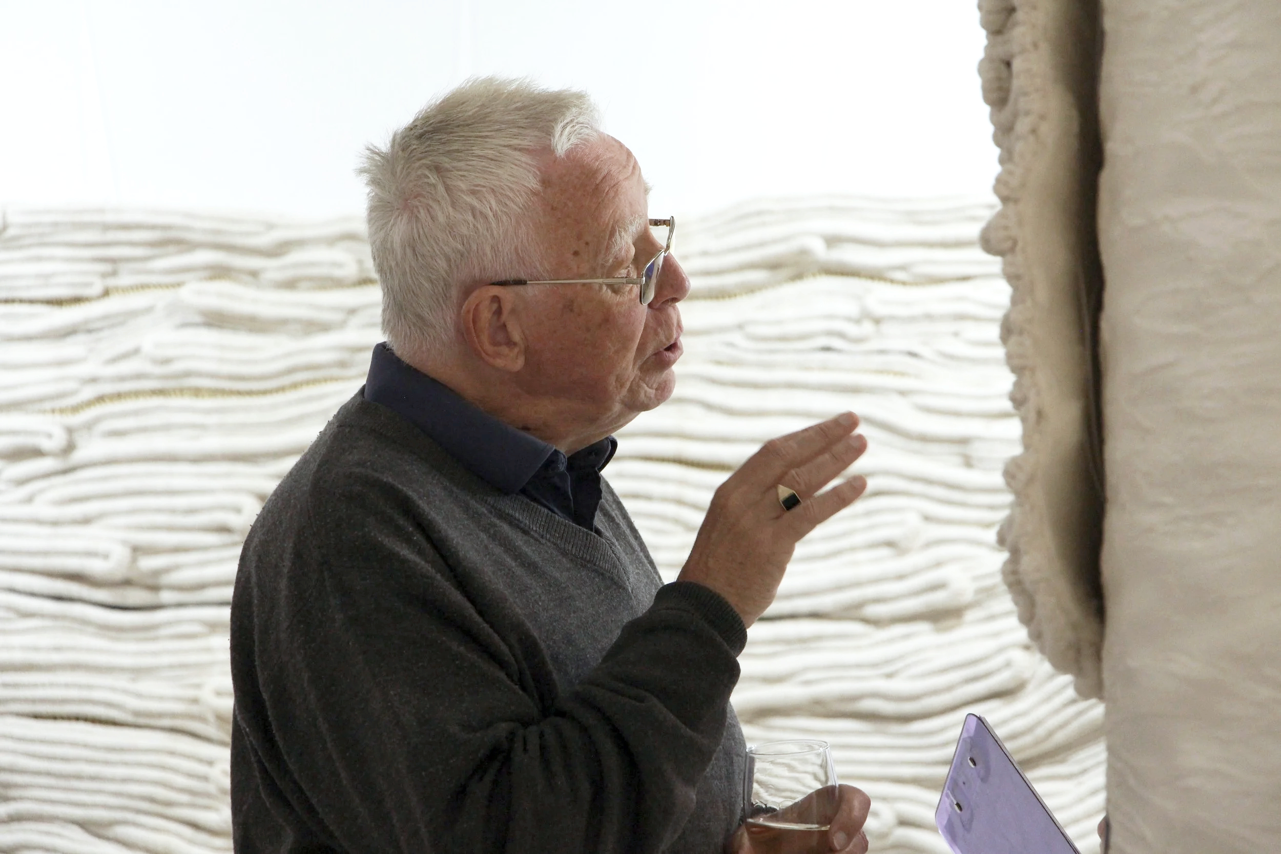 An elderly man with white hair, glasses, and wearing a dark sweater is holding a glass of wine and gesturing with his hand while talking at an art gallery.
