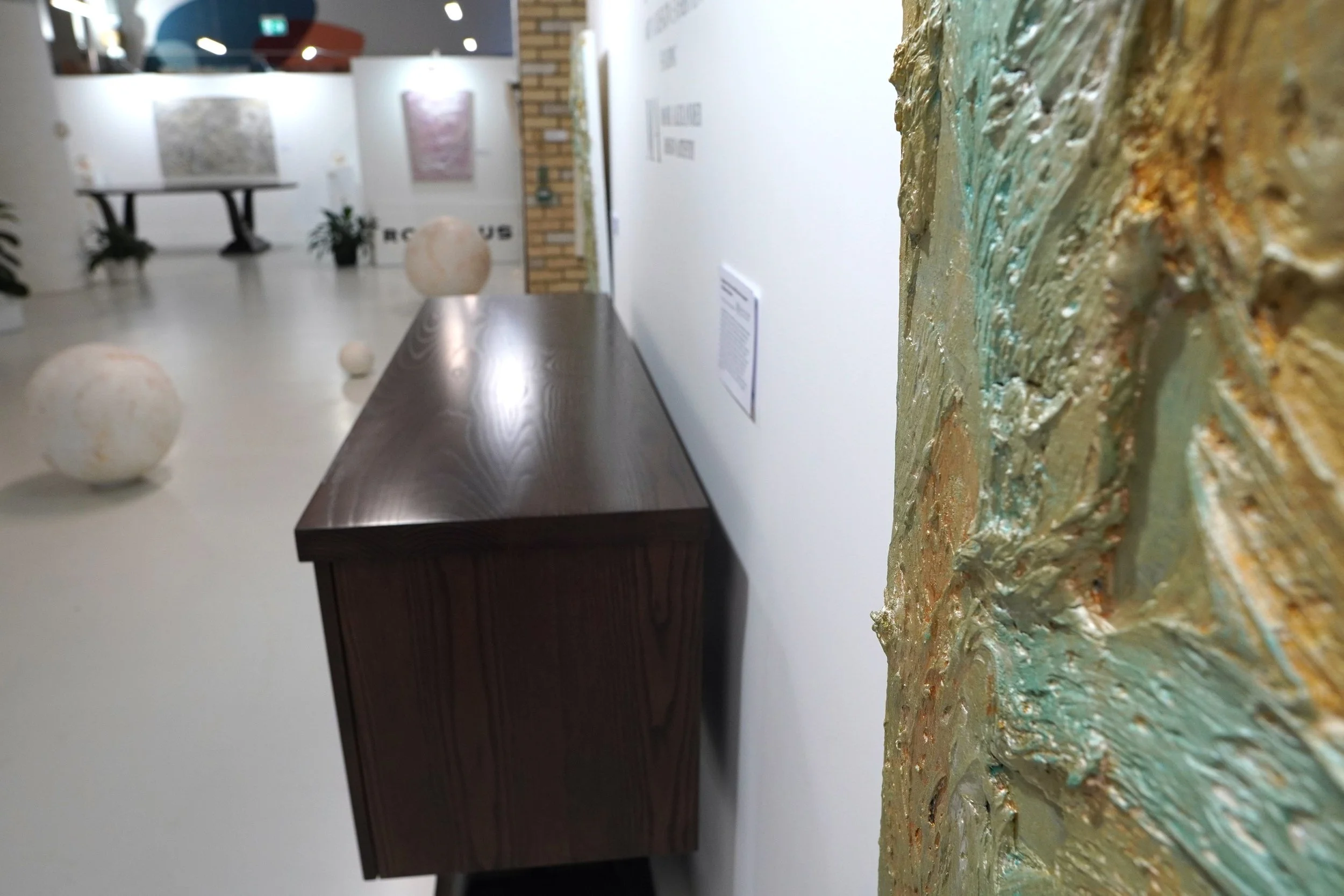 Art gallery with textured abstract paintings on the walls, a wooden display case, and spherical sculptures on the floor.