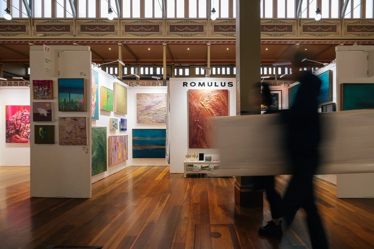 An art gallery with colorful paintings on white walls and wooden floors, two blurred people walking, and the sign 'ROMULUS' above one painting.