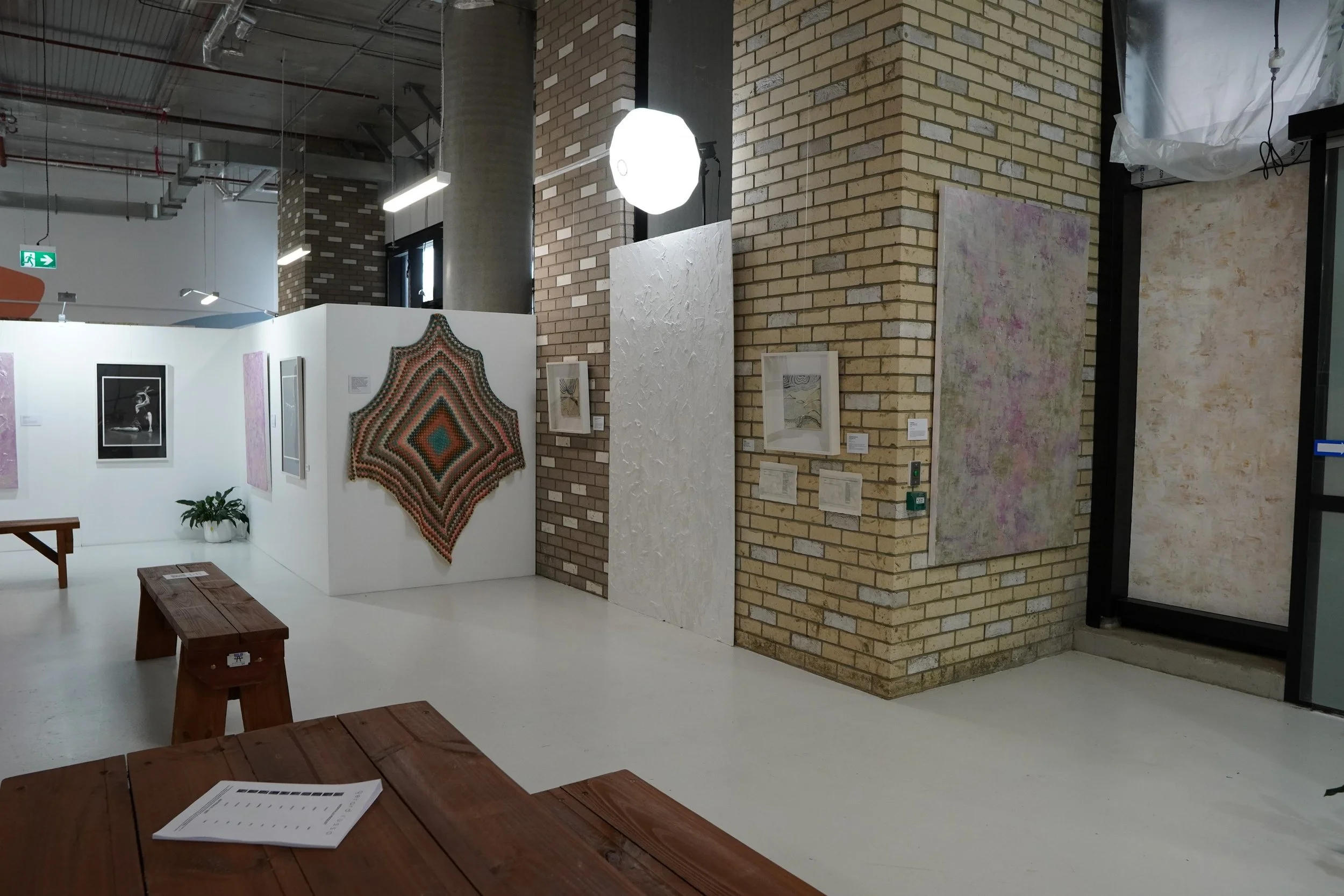 Art gallery with various artworks on display, including paintings and a textile piece, with wooden benches and a potted plant in a well-lit space with brick walls and industrial ceiling.