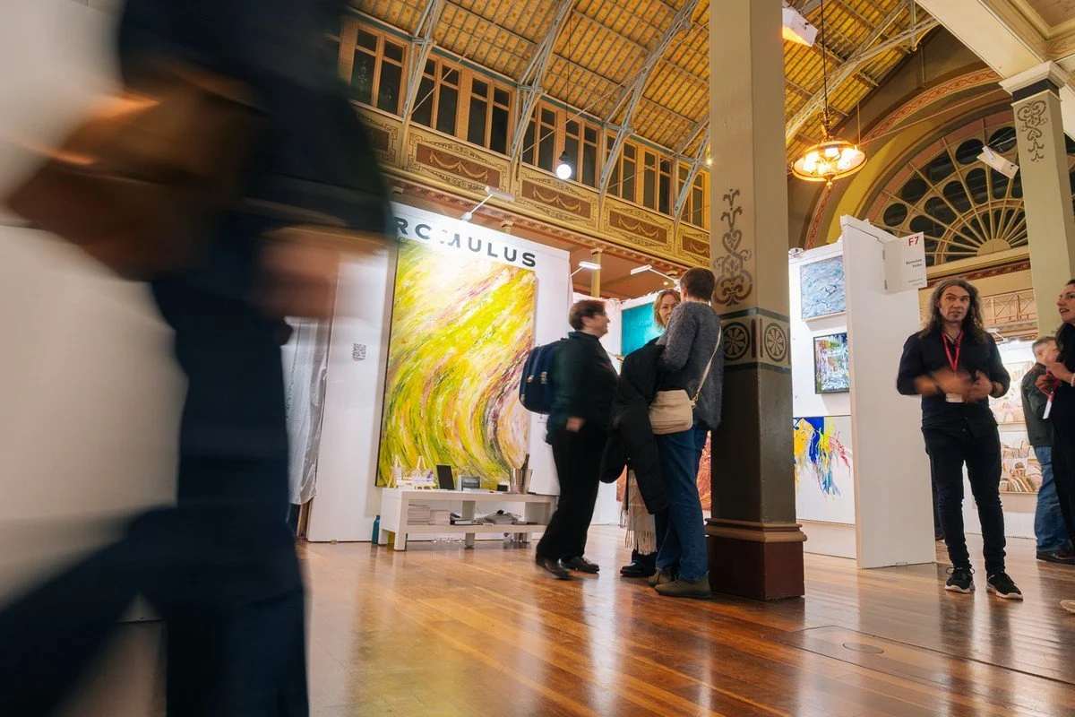 People attending an art exhibition inside a historic building with ornate decorations and high ceilings. They are viewing and discussing various colorful paintings, with some individuals standing close together in groups.