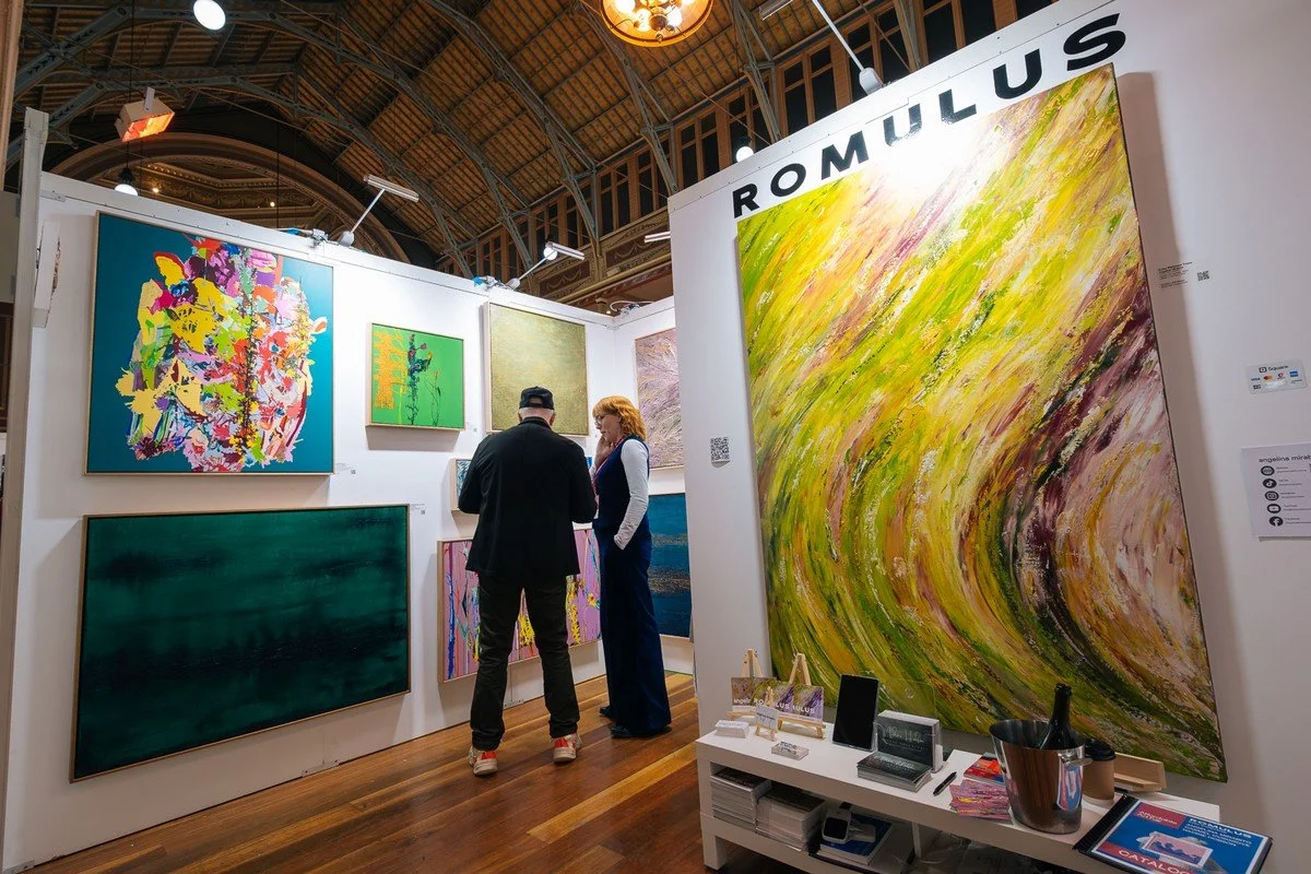Two people are in an art gallery standing in front of a white wall filled with colorful abstract paintings. A large yellow and green abstract artwork titled "ROMULUS" hangs prominently on the right. The gallery has a high arched ceiling with exposed 