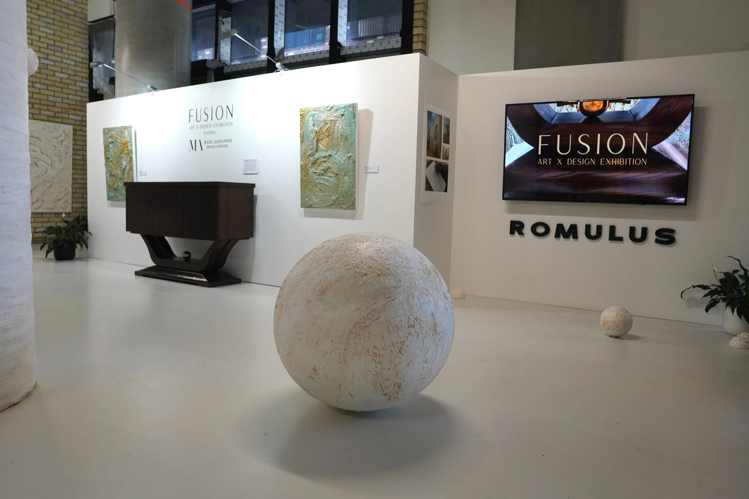 Art display at an exhibition featuring a large white textured sphere in the foreground, a dark wooden cabinet in the background, and a white wall with two abstract paintings, the words 'FUSION ART X DESIGN EXHIBITION,' and a flat screen television di