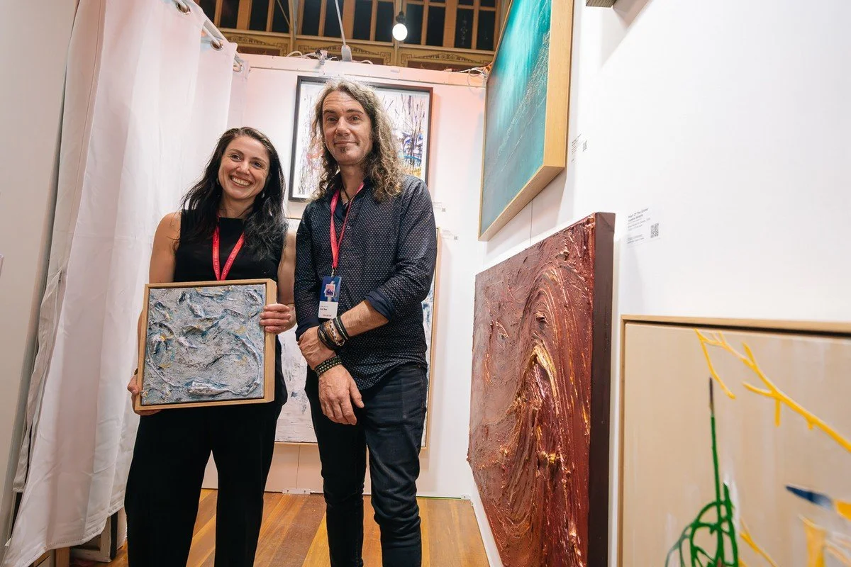 Two people, a woman and a man, standing in an art gallery. The woman is holding a textured abstract painting and smiling at the camera. The gallery's walls display various colorful artworks.