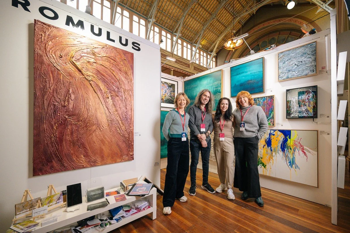 Four people standing in front of an art exhibit at an art gallery, with various colorful paintings displayed on the walls behind them.