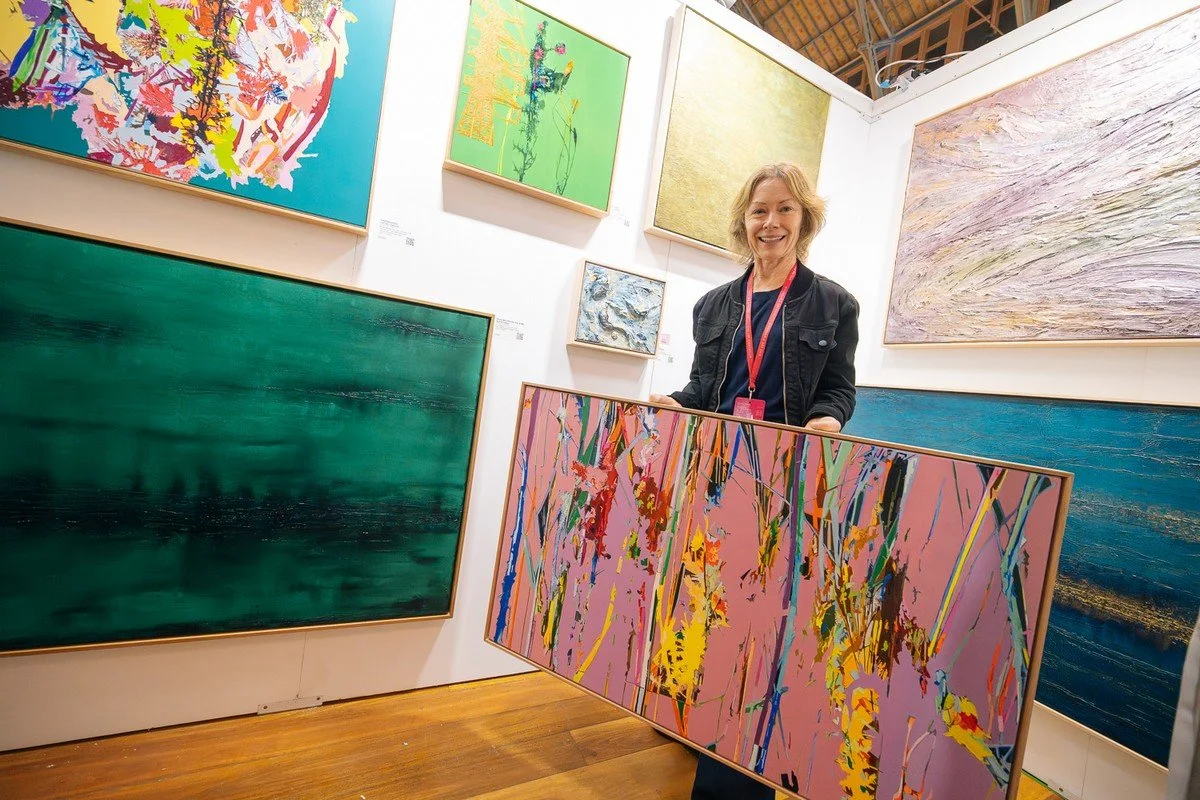 A woman holding a colorful abstract painting in an art gallery with various other paintings on the walls.