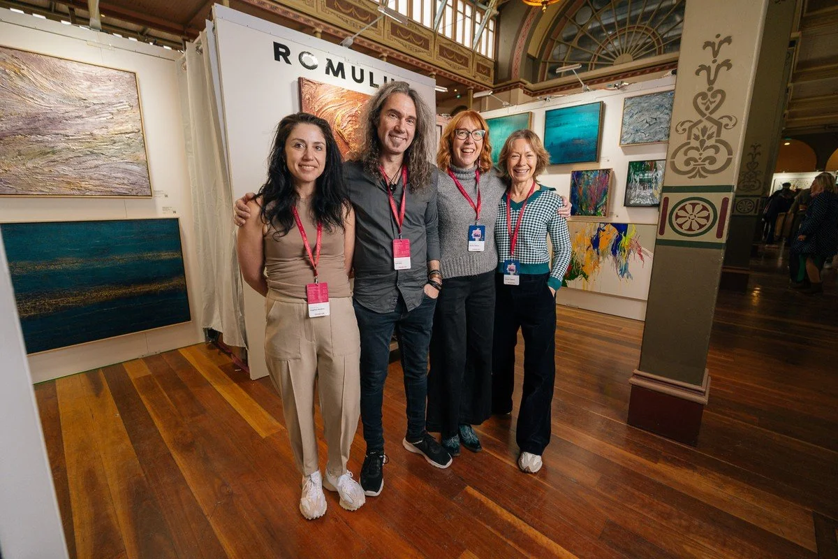 Four women standing together at an art exhibition, smiling. They are in front of a display of colorful paintings and a sign that reads 'ROMULLI'. They are wearing name tags on lanyards around their necks. The gallery has wooden floors and decorative 