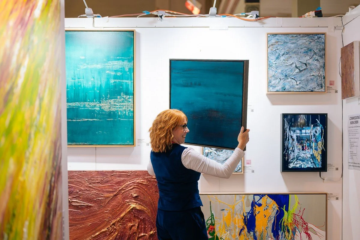 A woman with reddish hair and glasses hanging in an art gallery, taking a photo of a blue abstract painting on the wall surrounded by various other colorful artworks.