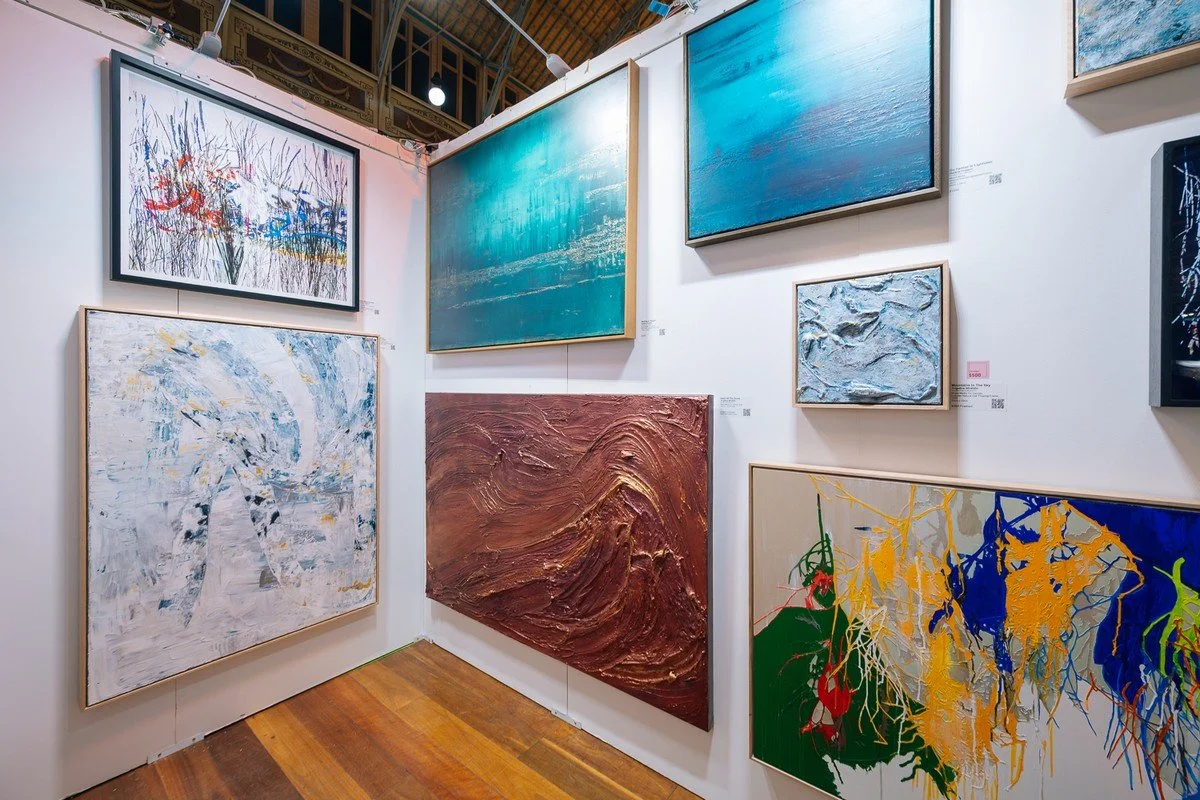 A gallery wall with various abstract and landscape paintings in different sizes and colors, including blue, white, brown, and multicolored artworks.