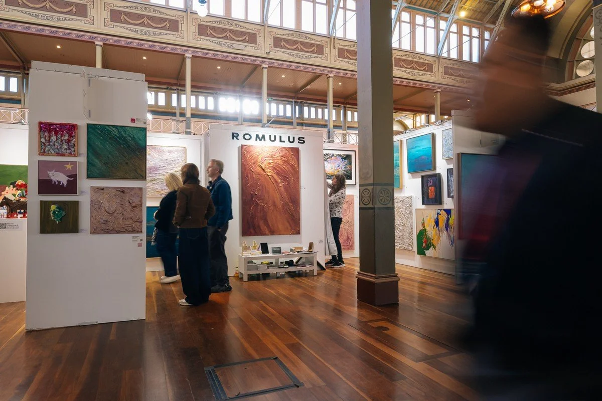 An art gallery with abstract paintings displayed on white walls, visitors viewing the artwork, and a large sign that reads 'ROMULUS' on one of the walls.