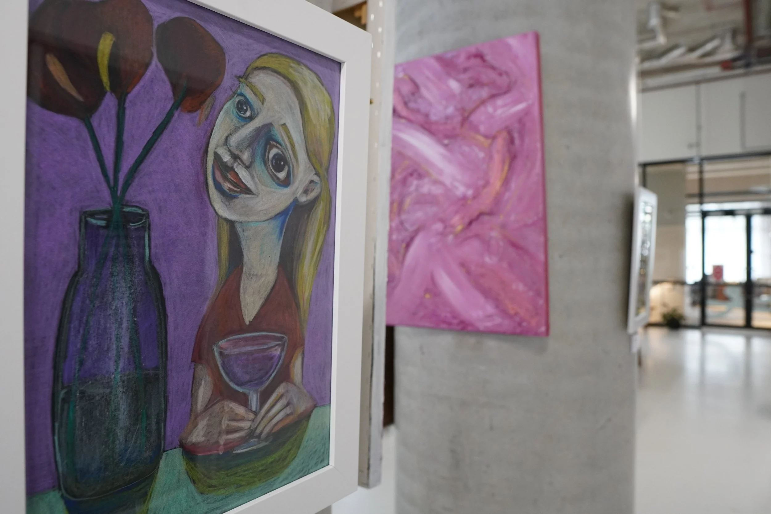 Colorful painting of a woman with exaggerated facial features, blonde hair, and large eyes, holding a glass of wine with a vase of flowers in the background, displayed in a gallery with pink abstract art and a modern interior.