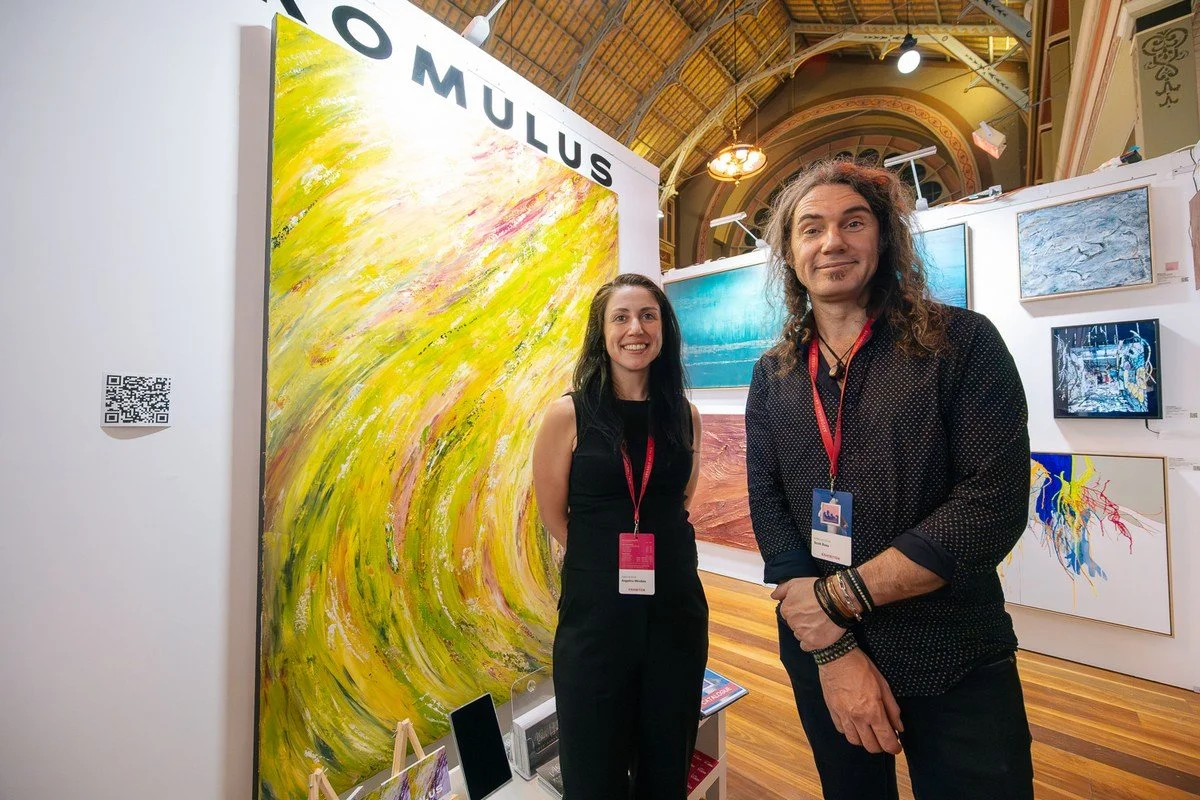 Two smiling women standing in an art gallery or exhibition space with paintings on the wall behind them and a large abstract yellow artwork to their left.