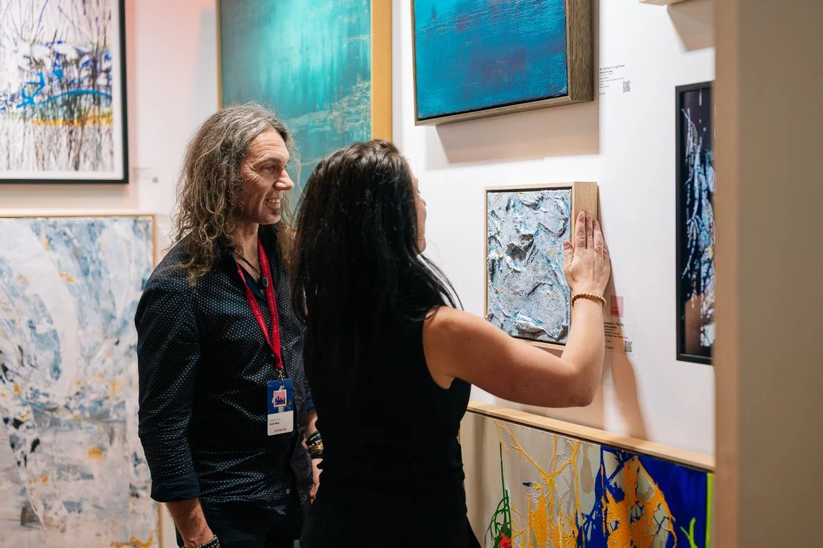 Two women are viewing and discussing artwork at an art gallery. One woman is touching a textured painting on the wall, and the other is looking on. Several other paintings are displayed on the walls and in the background.