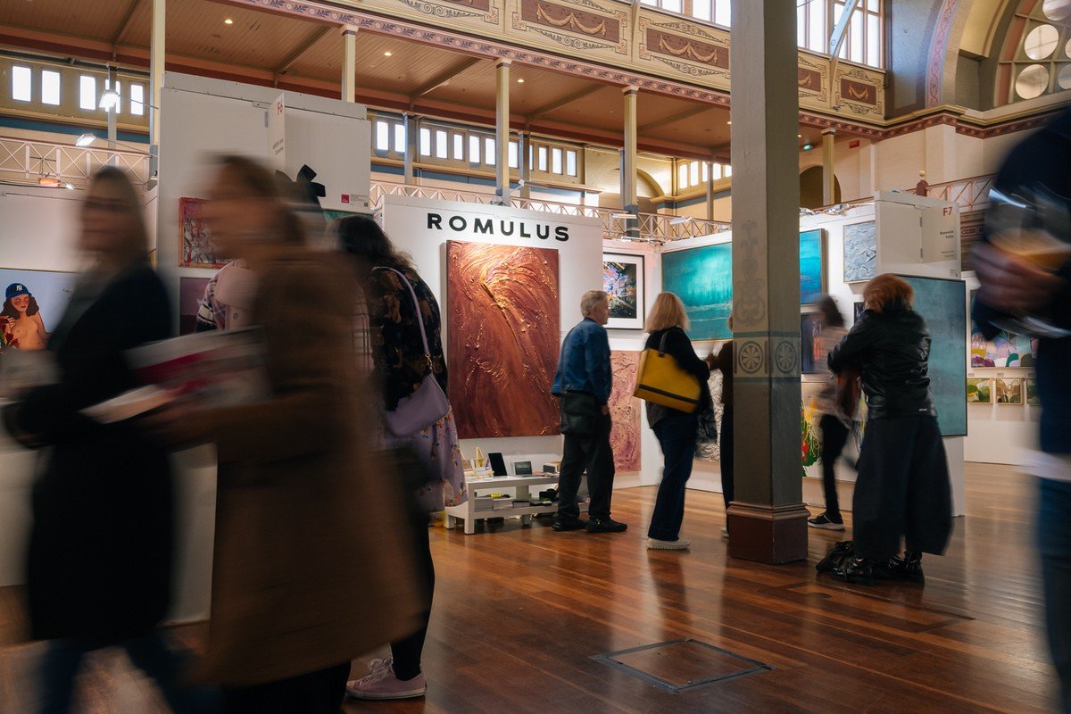 An art gallery with multiple abstract paintings displayed on white walls. Several visitors walk and look at the artwork, and the gallery has a high ceiling with decorative moldings and large windows.