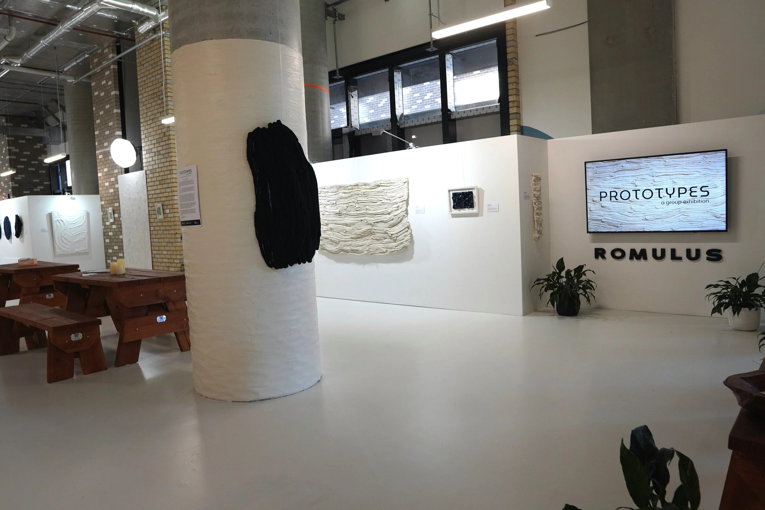 An art exhibition display with sculptures and framed artworks on white walls, featuring a digital screen with the text 'PROTOTYPES a group exhibition' and the names 'ROMULUS'. The setting has potted plants and wooden benches.