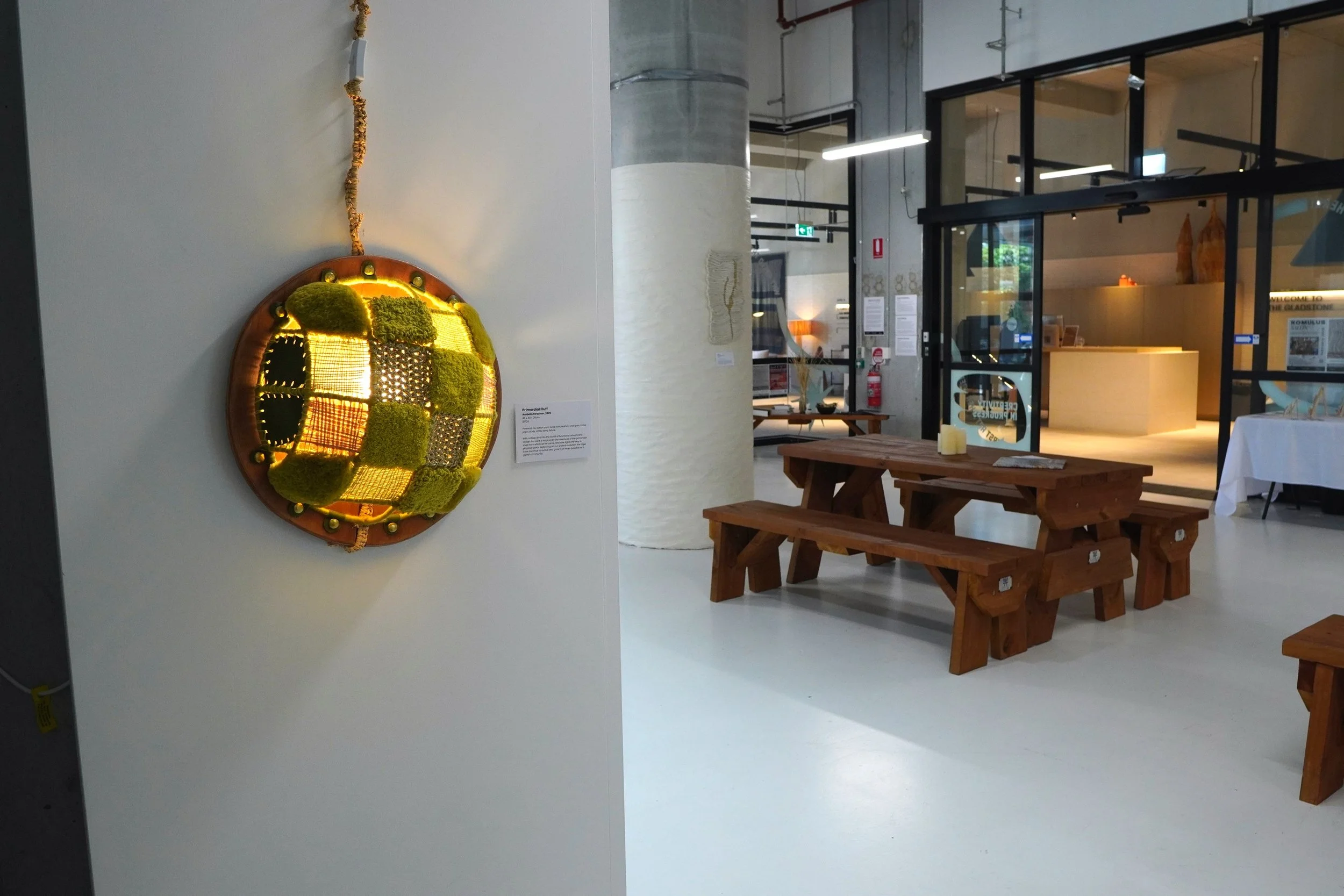 An art piece on a white wall composed of squares and rectangles of moss and other textured materials, with backlighting. The photo shows inside a gallery with wooden benches, a white display table, and an industrial-style interior with exposed pipes 
