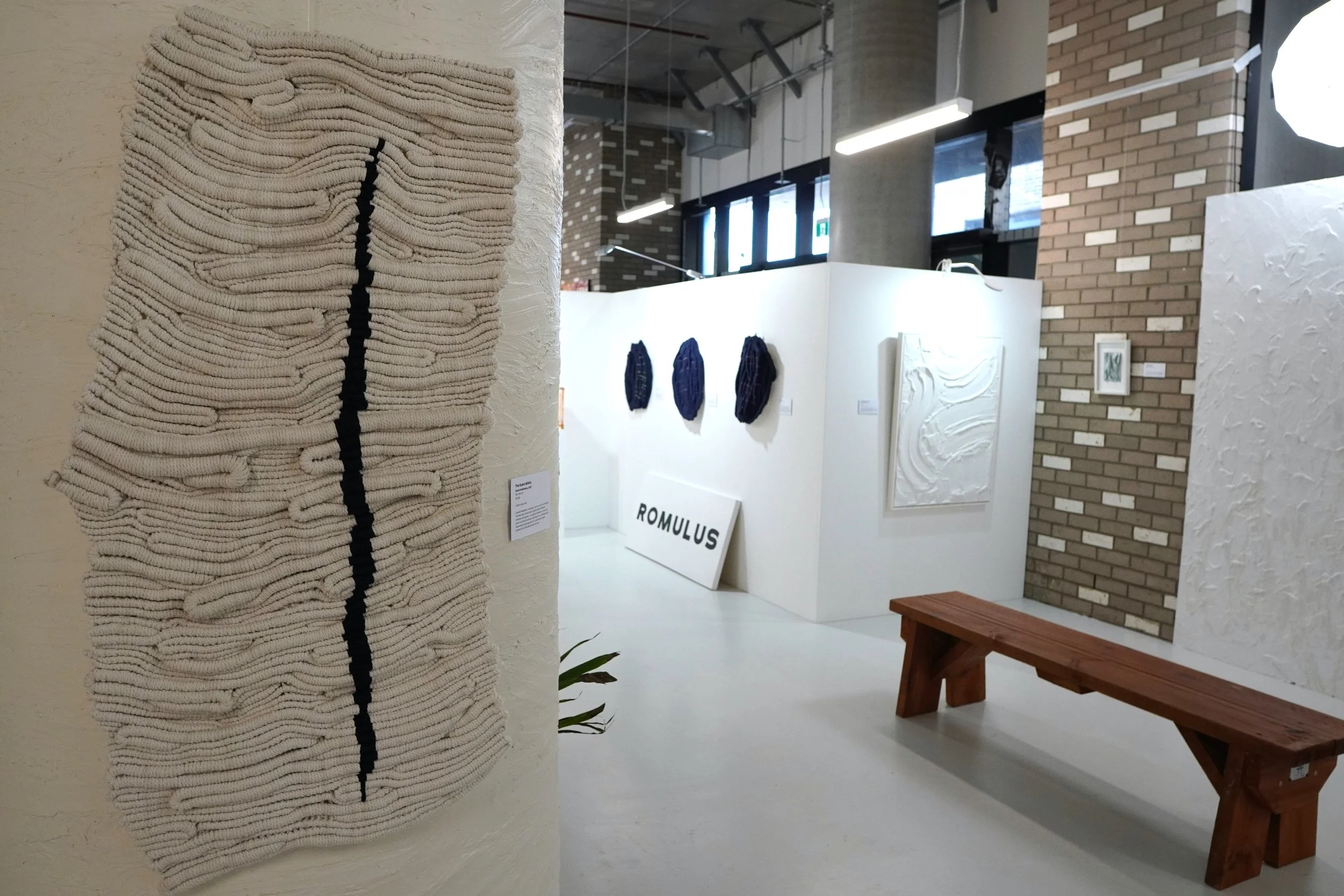 An art gallery with textured woven wall art on a white pillar, paintings or textiles on a white wall, a wooden bench, and a brick wall. The display includes a sign reading 'ROMULUS'.