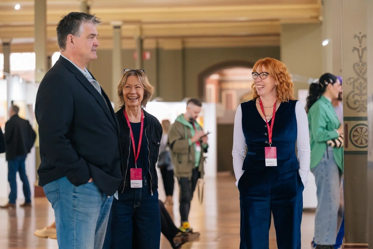 Three people, a man and two women, are standing and talking at an indoor event. They are smiling, and all are wearing lanyards with badges.